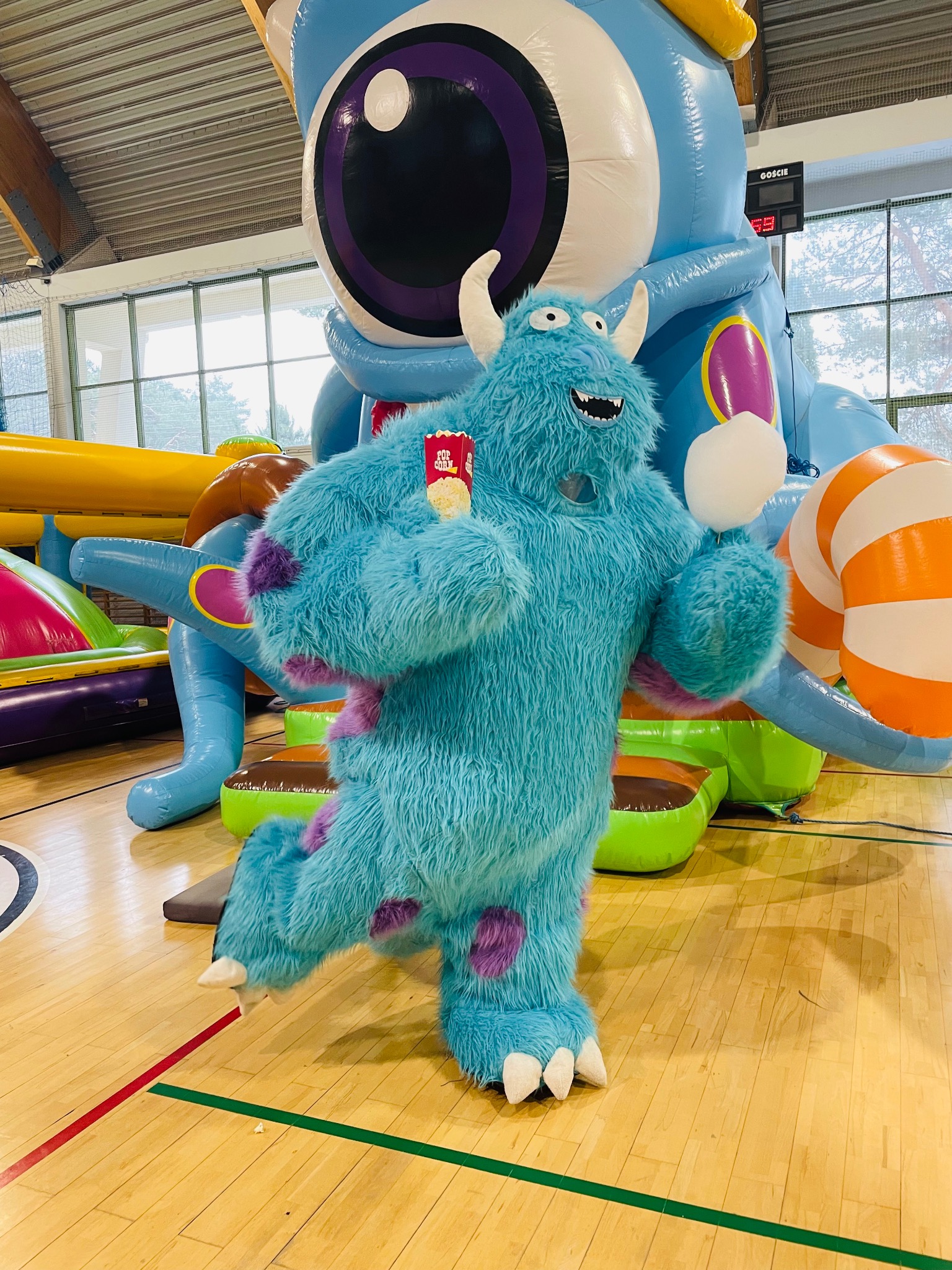 Animator w kostiumie Sully z Monsters, Inc. z popcornem, na tle dmuchańca w Łodzi. Atrakcja eventowa dla dzieci z unikalnym strojem i scenografią.