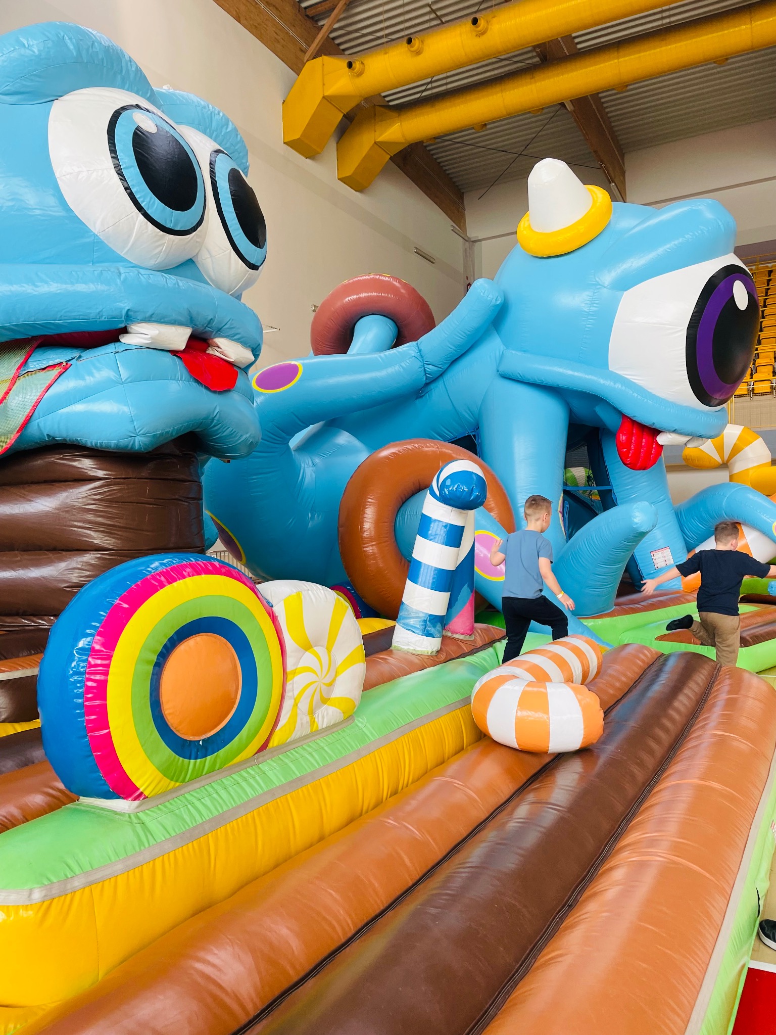 Dzieci bawią się na dmuchanym placu zabaw z kolorowymi przeszkodami i postaciami, w tym niebieskimi potworami z wystającymi językami i dużymi oczami.