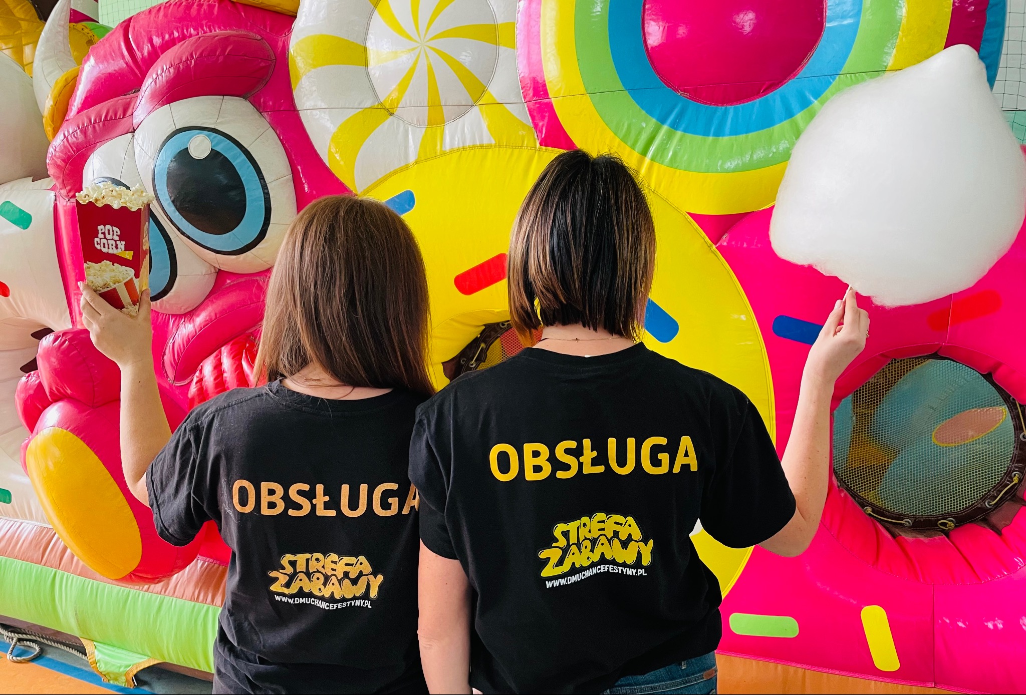 Obsługa Strefy Zabawy w czarnych koszulkach z popcornem i watą cukrową na tle kolorowego dmuchańca, z logo firmy widocznym na koszulkach.