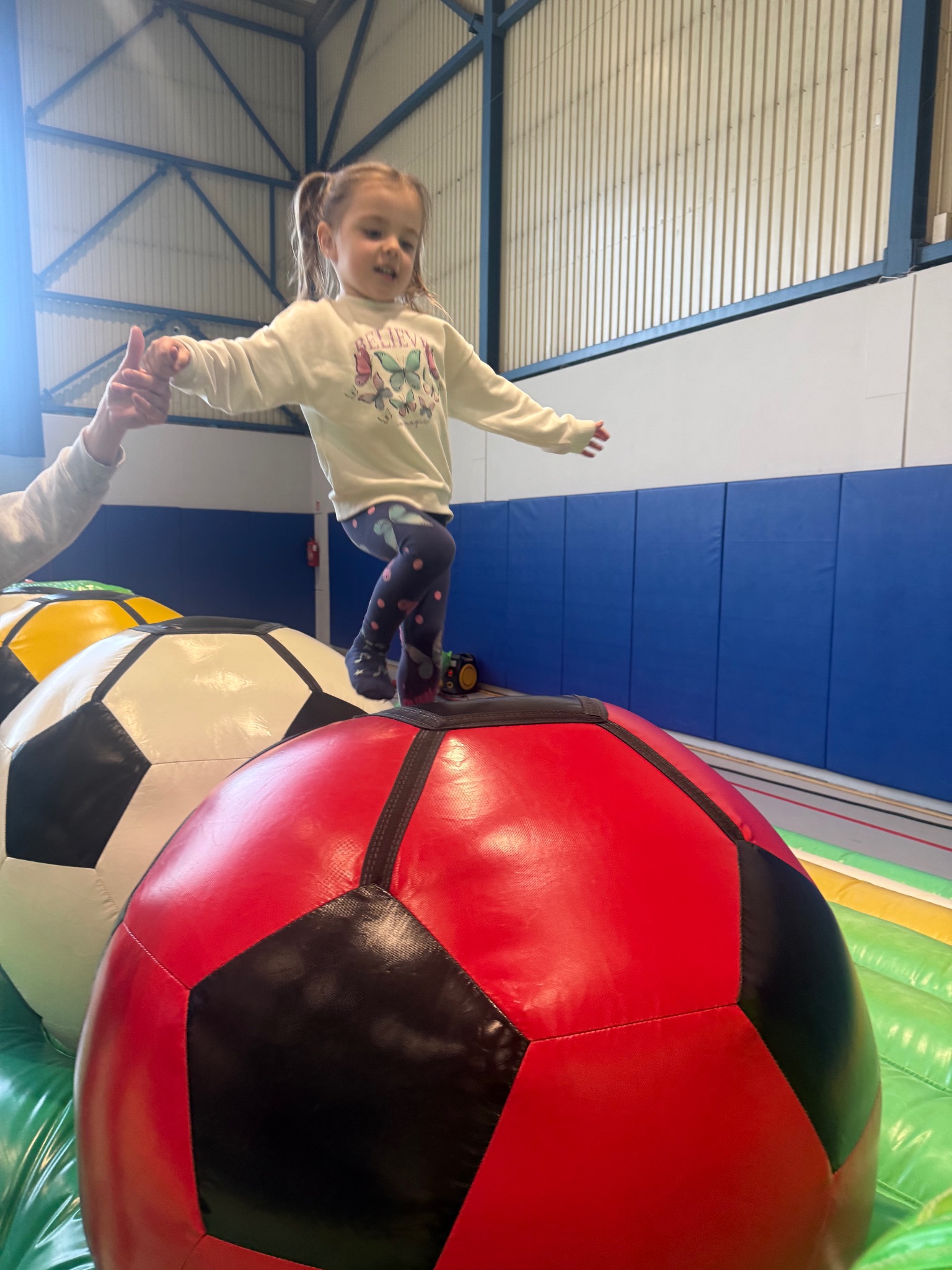 Dziewczynka skacze po dmuchanych piłkach na zielonym materacu. Dłoń osoby dorosłej asekuruje dziecko. Arena zabaw w hali.