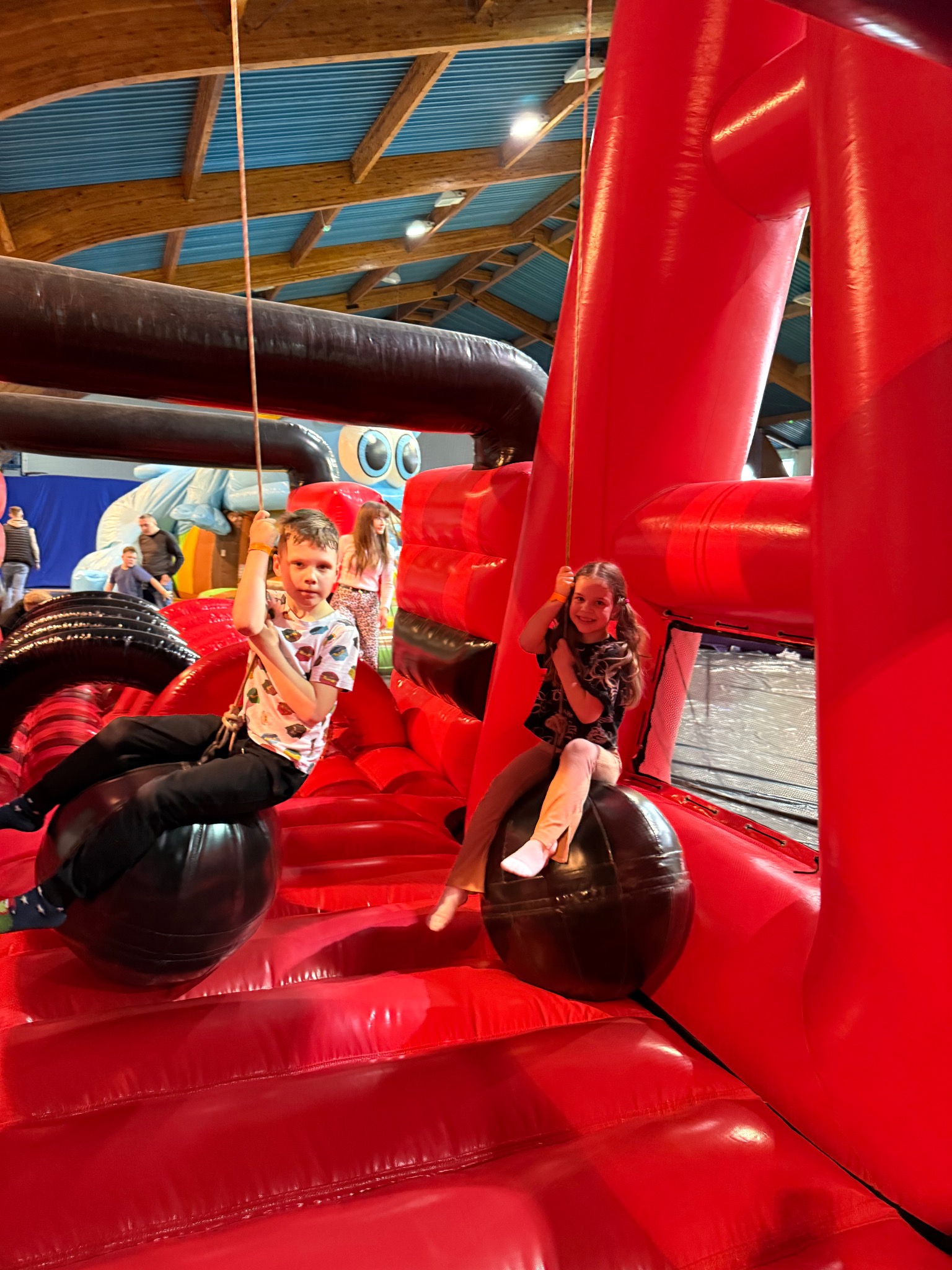 Dwoje dzieci huśta się na czarnych kulach w czerwonym dmuchanym parku rozrywki z niebieskim sufitem i drewnianymi belkami w tle.