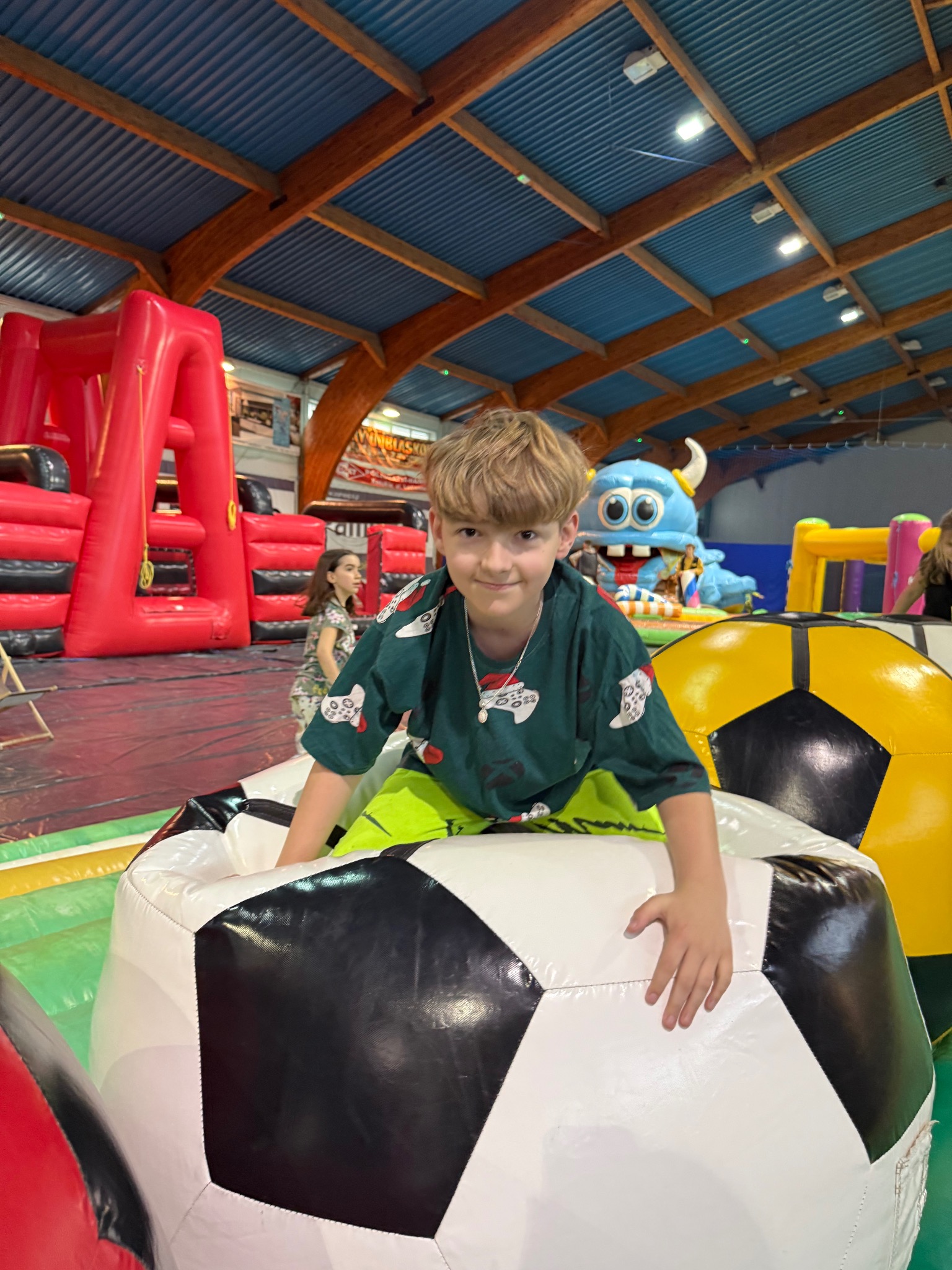 Chłopiec w koszulce z motywem gier w dmuchanym basenie z piłkami, w tle dmuchane konstrukcje i inne dzieci w sali zabaw.