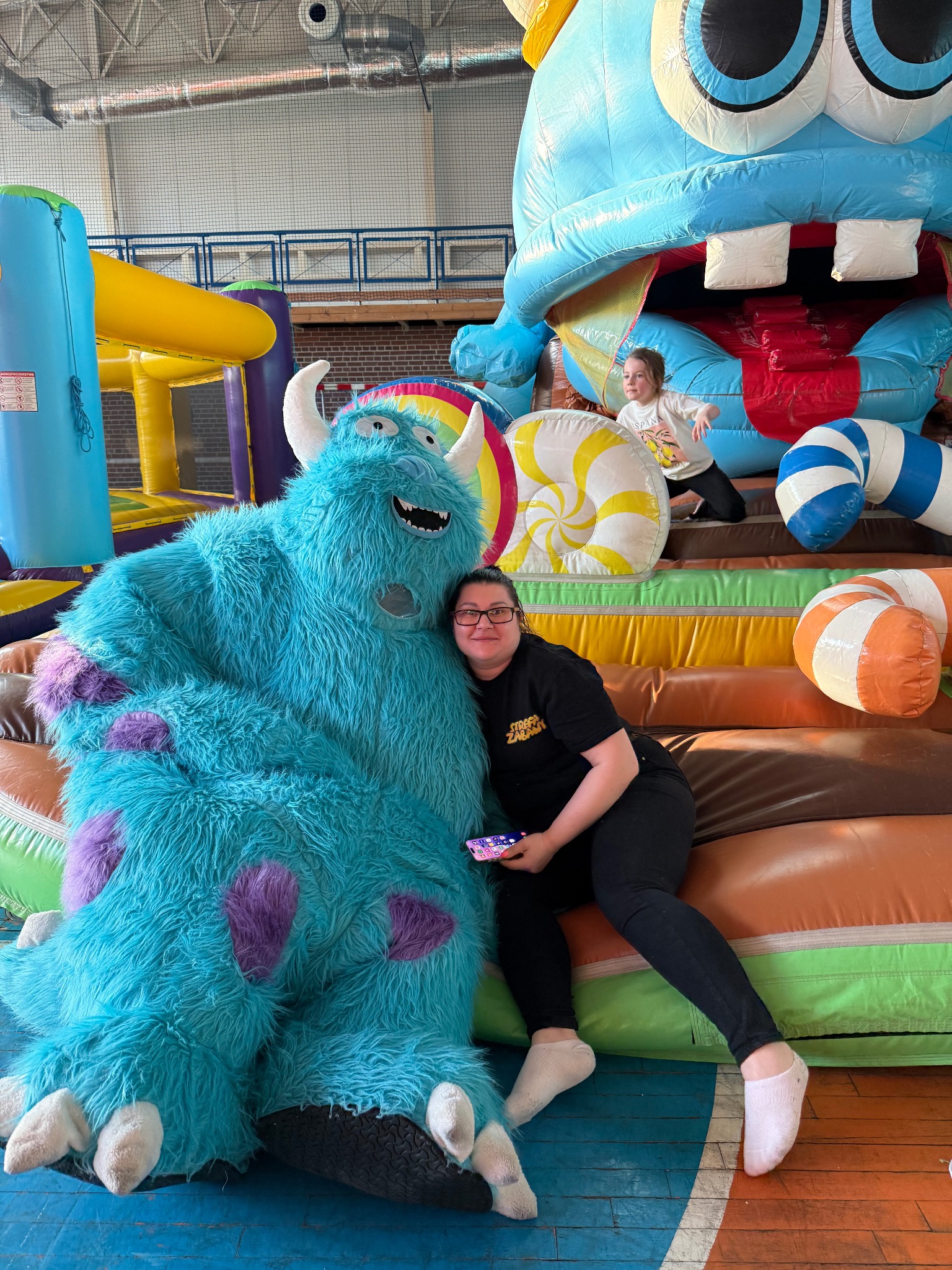 Kobieta obejmuje postać Sully z Potworów i Spółki na dmuchanym zamku; w tle dziecko na dmuchańcach, atrakcja imprezowa.