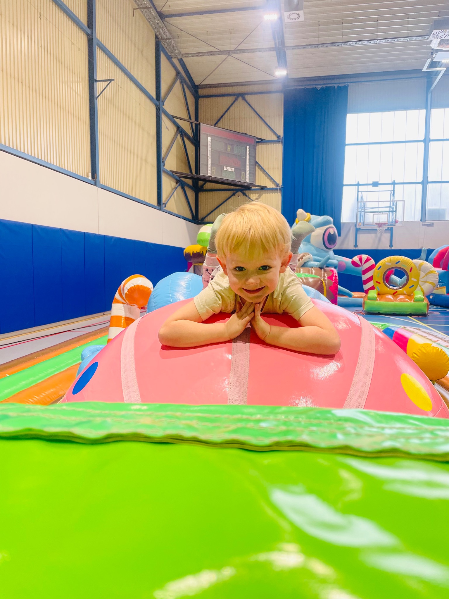 Uśmiechnięty chłopiec leży na różowym dmuchańcu w hali sportowej z innymi kolorowymi dmuchańcami w tle, tworząc radosną scenę.