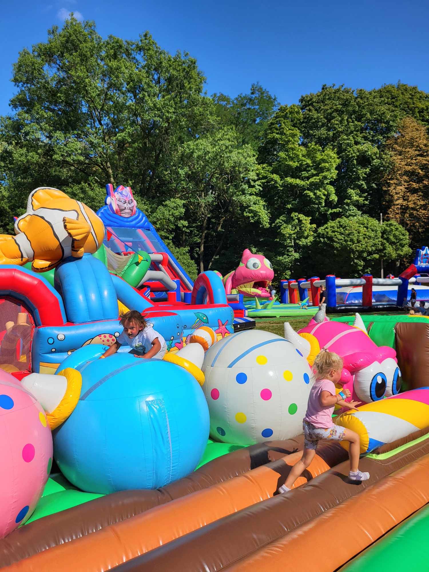 Dzieci bawią się na dmuchanym placu zabaw z motywami zwierząt i postaci z bajek, w tle drzewa i niebieskie niebo. Kolorowe atrakcje dla dzieci na imprezie plenerowej.