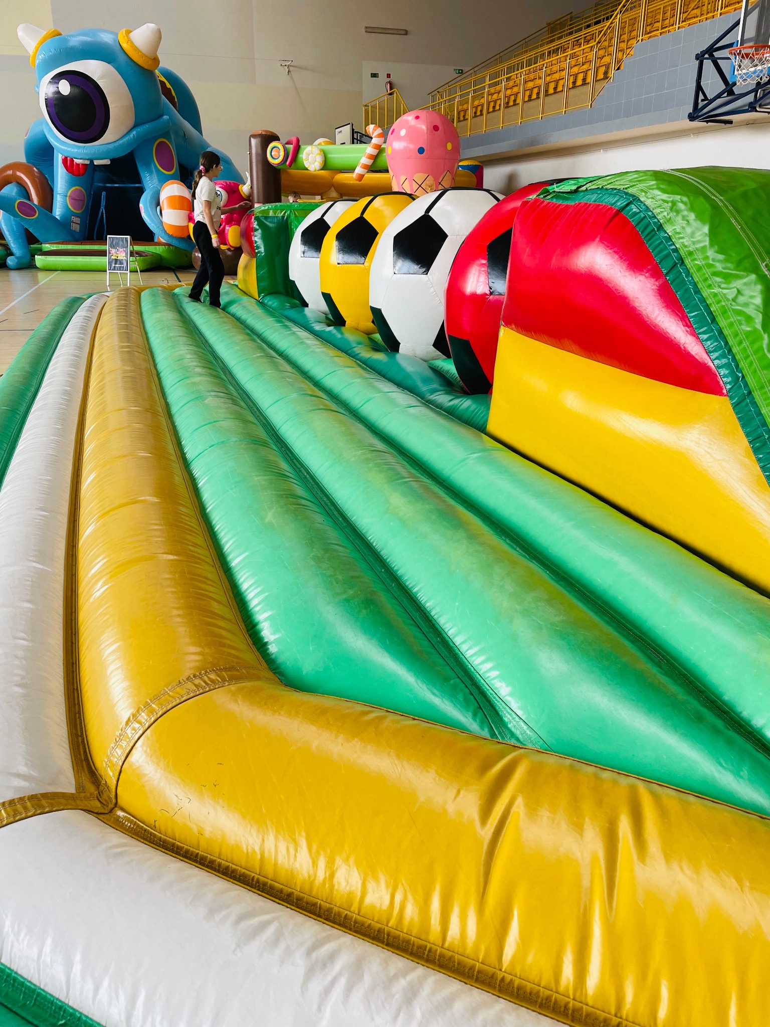 Kolorowy dmuchany tor przeszkód z motywem piłek i postaciami z bajek w hali sportowej. Dziecko wchodzi na atrakcję. Widoczne trybuny.