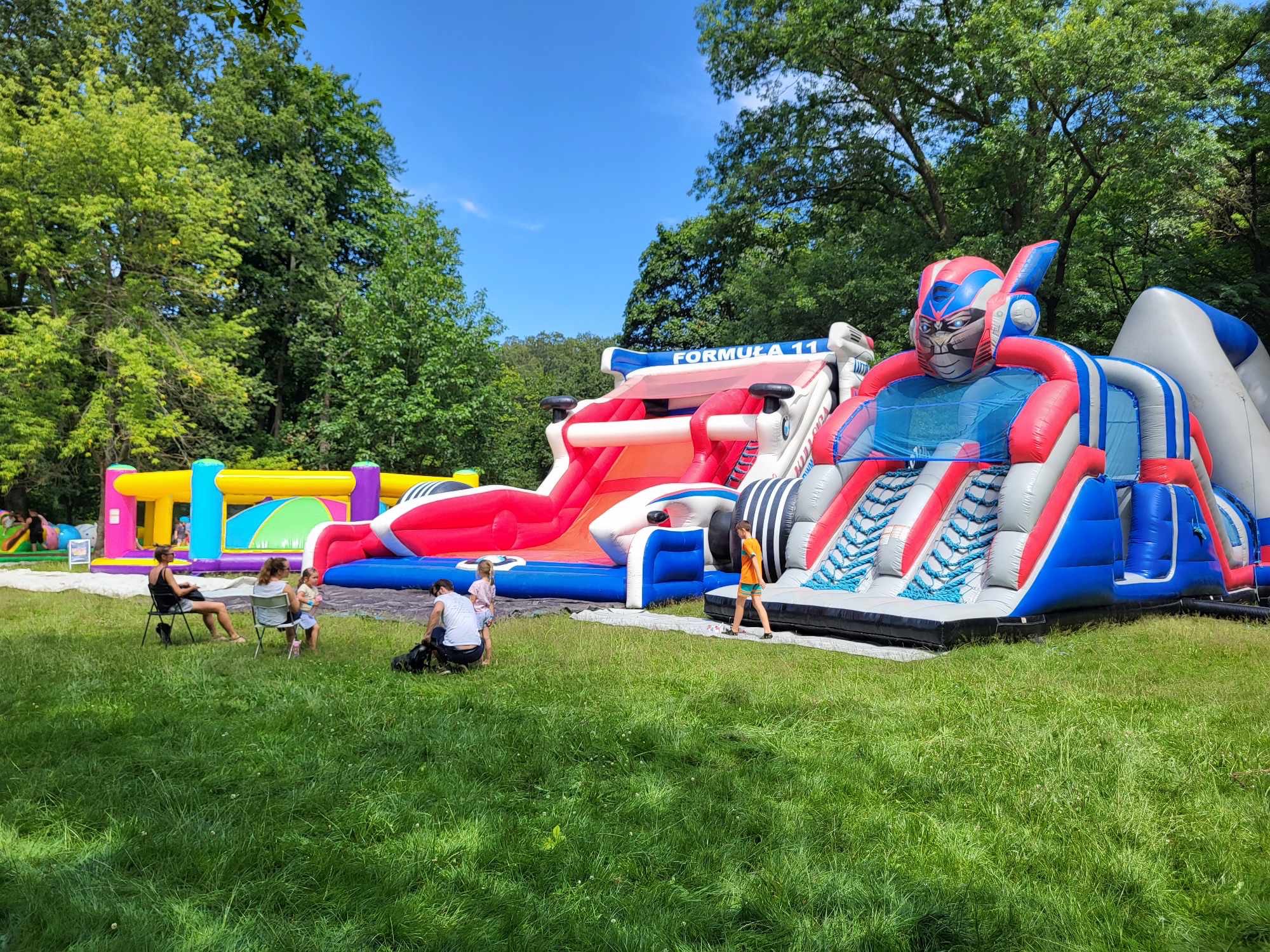 Dwa dmuchańce: tor przeszkód z motywem Formuły 1 i zjeżdżalnia w kształcie robota, na trawie, w tle drzewa i niebieskie niebo. Dzieci i dorośli w pobliżu.