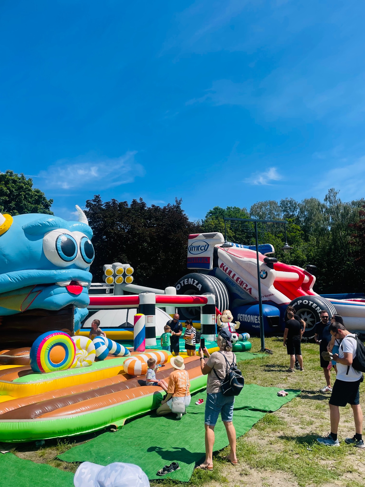 Kolorowy plac zabaw dmuchanych dla dzieci z motywami bajkowymi i sportowymi, otoczony zielenią i bawiącymi się osobami w słoneczny dzień.