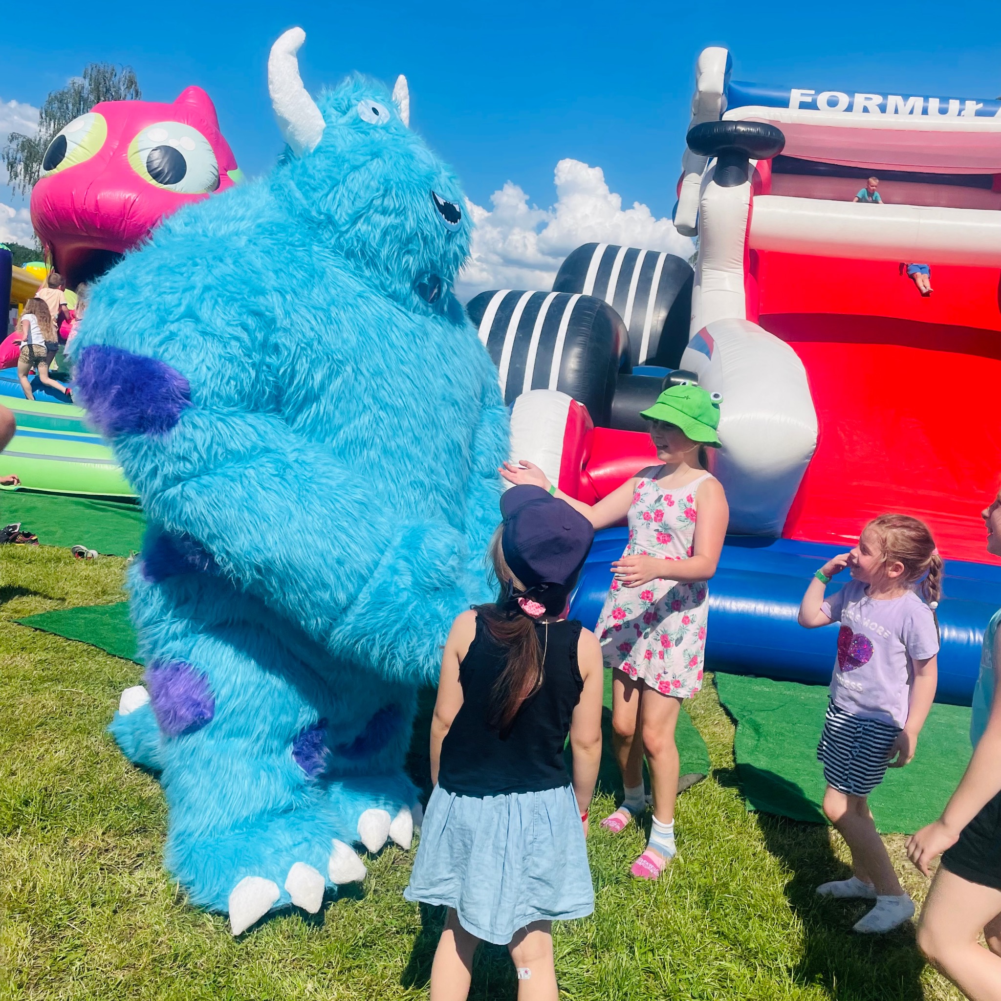 Dzieci przy dmuchanym zamku i pluszowym Sully na zielonej trawie w słoneczny dzień. Zamek ma motyw wyścigowy z napisem 'Formula'.