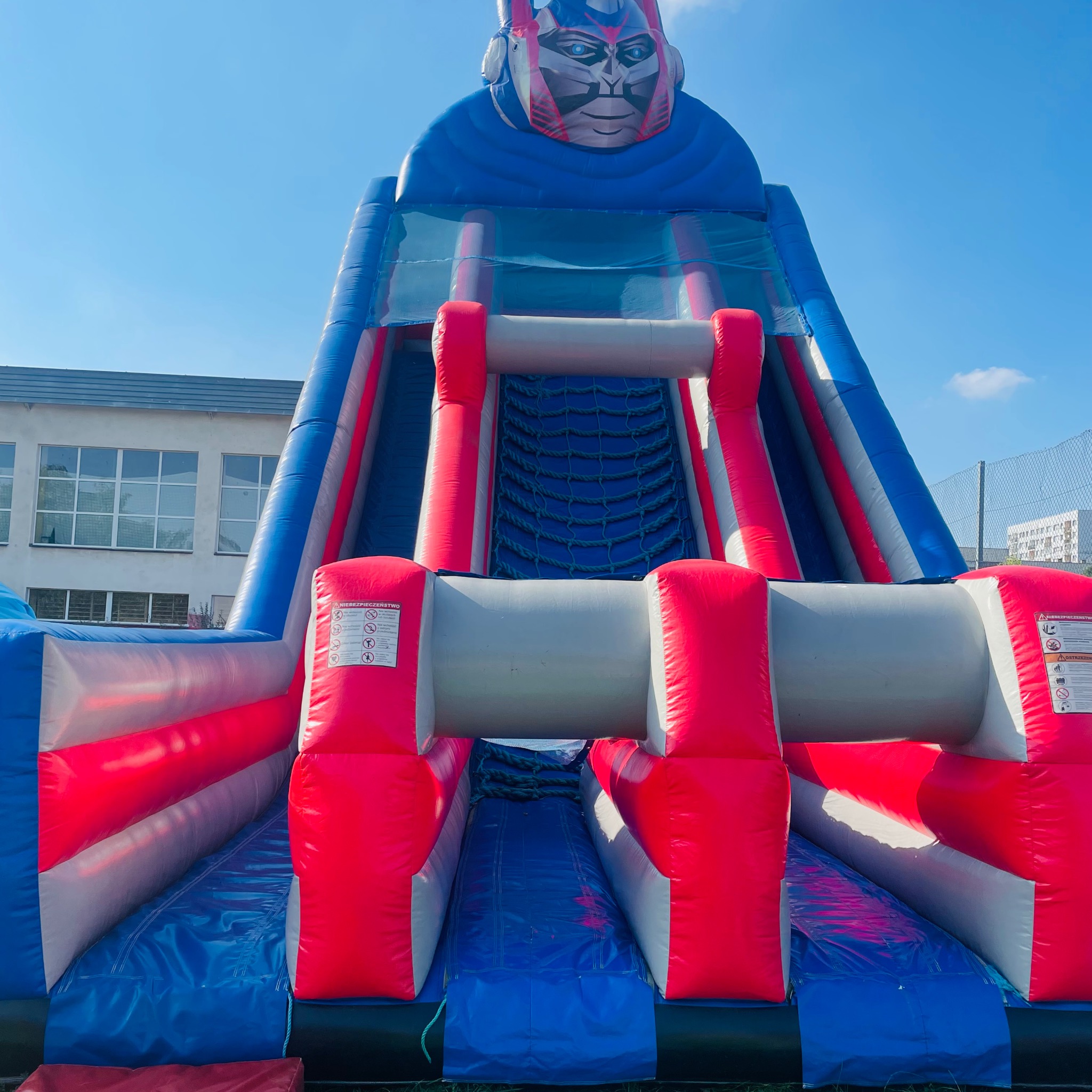 Niski kąt na dmuchaną zjeżdżalnię z motywem robota, z niebiesko-czerwonymi elementami, stojącą na trawie, z budynkiem w tle i błękitnym niebem.