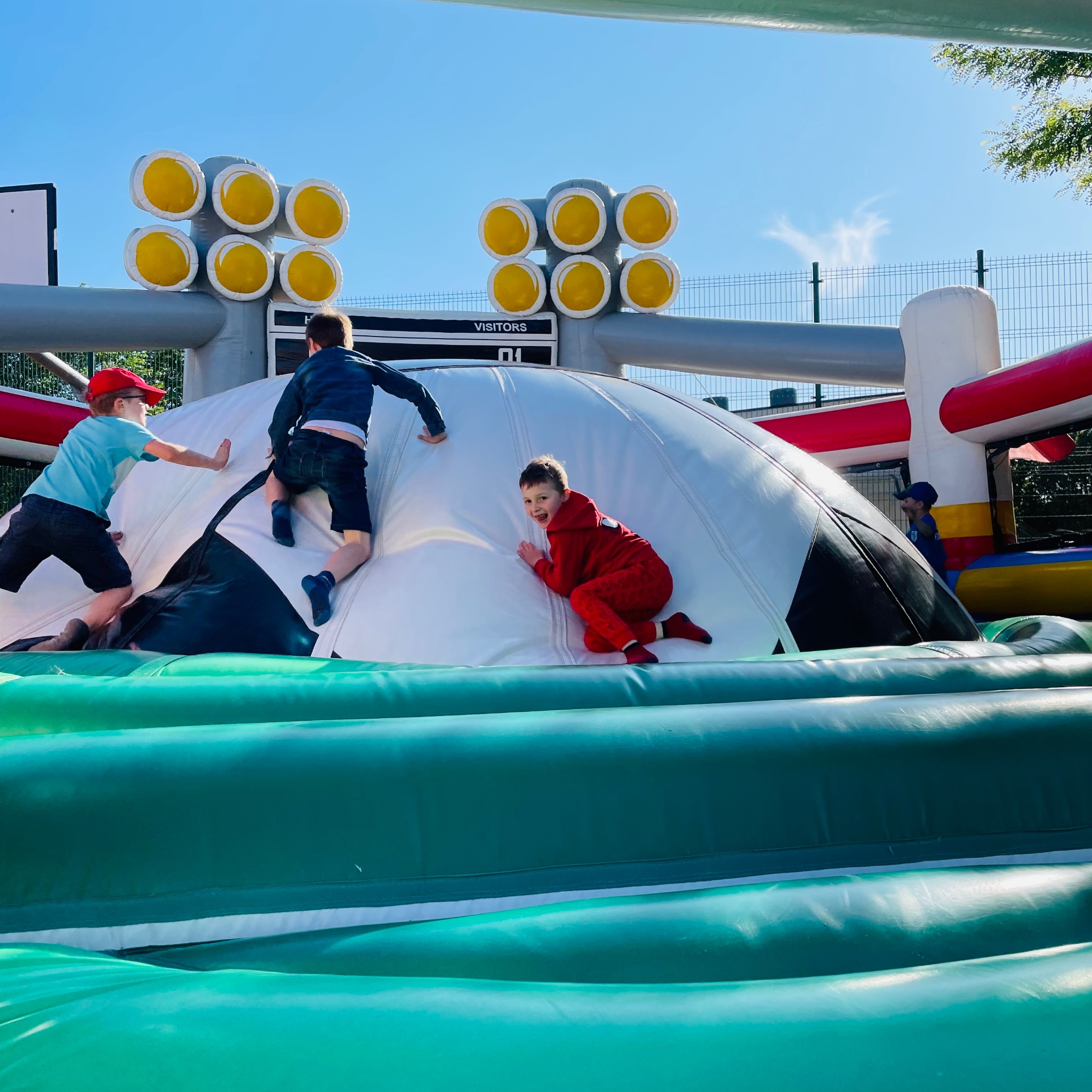 Dzieci bawią się na dmuchanym boisku piłkarskim z tablicą wyników i jaskrawymi reflektorami. Radosna zabawa na świeżym powietrzu w słoneczny dzień.