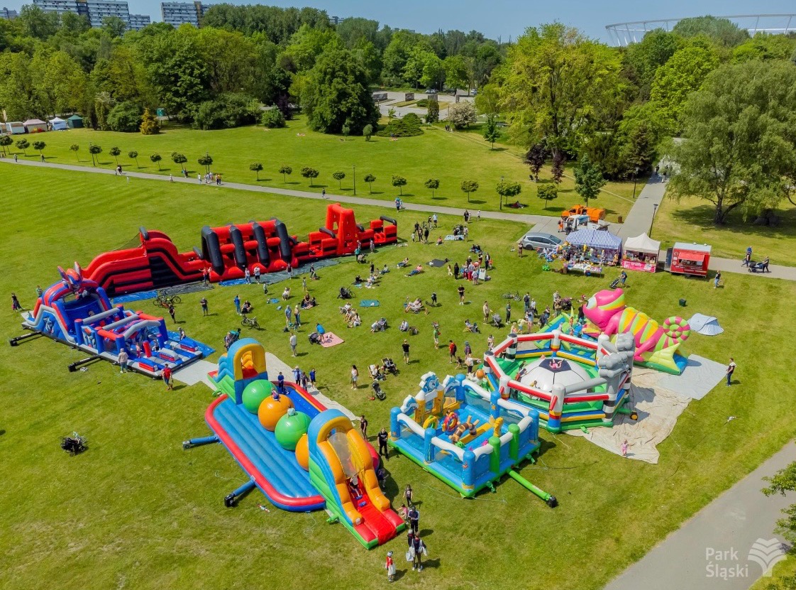 Park Śląski: Dmuchańce na zielonym trawniku, widok z góry. Różnorodne konstrukcje, w tym tor przeszkód i zjeżdżalnie, otoczone bawiącymi się ludźmi. Słoneczny dzień.