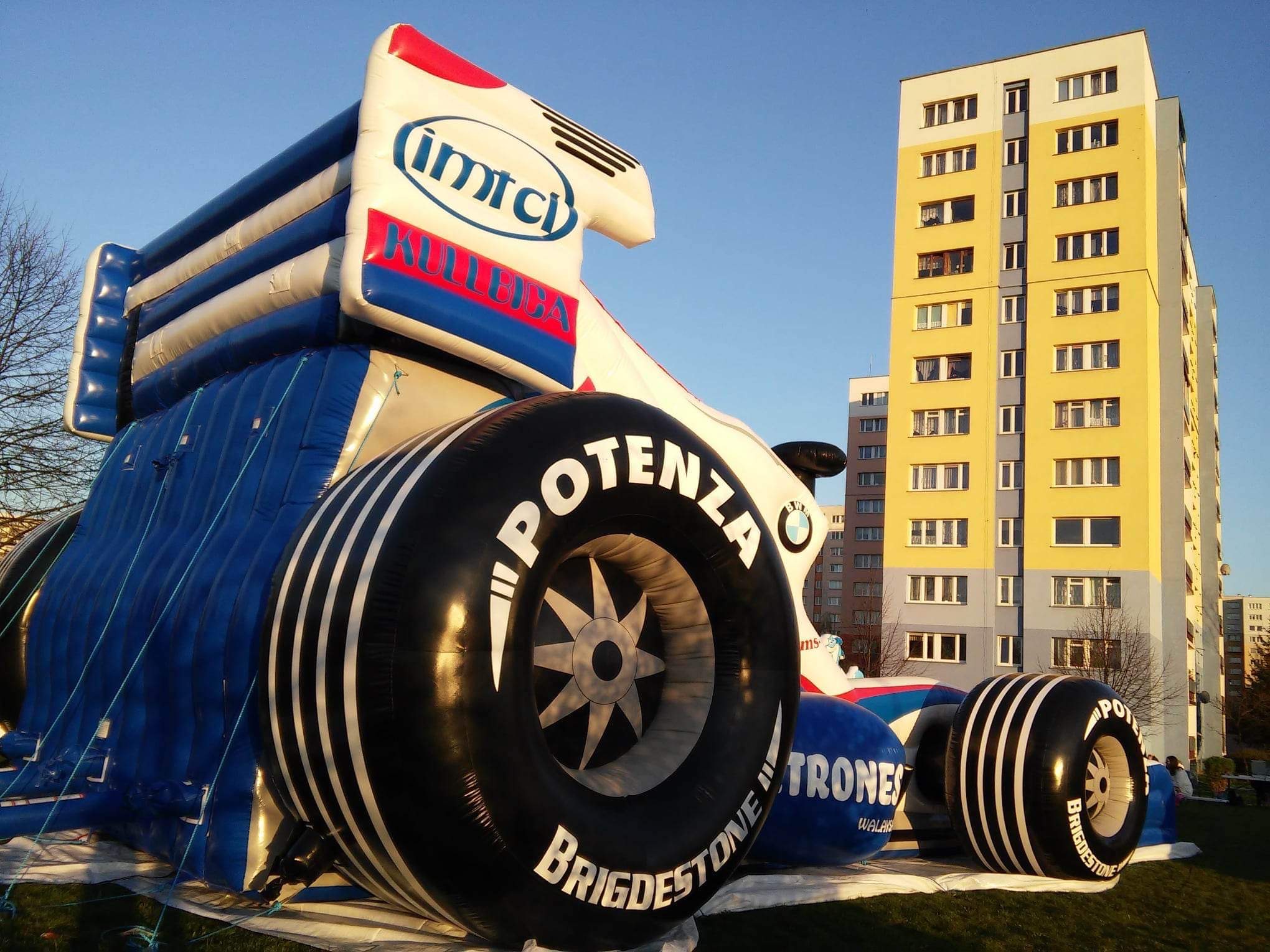 Nietypowy dmuchany bolid Formuły 1 z widocznymi oponami Bridgestone Potenza, ustawiony na trawie przed blokiem mieszkalnym w słoneczny dzień.