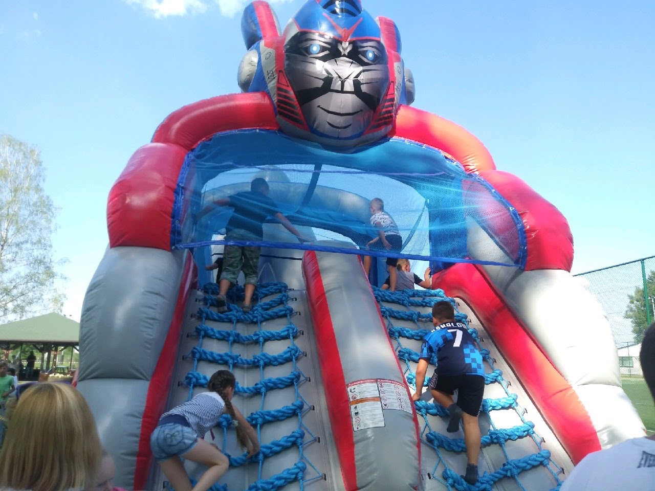 Dzieci wspinają się na dmuchany plac zabaw w kształcie robota z niebieskimi linami i siatką zabezpieczającą. Czerwono-szaro-niebieska kolorystyka, słoneczny dzień.