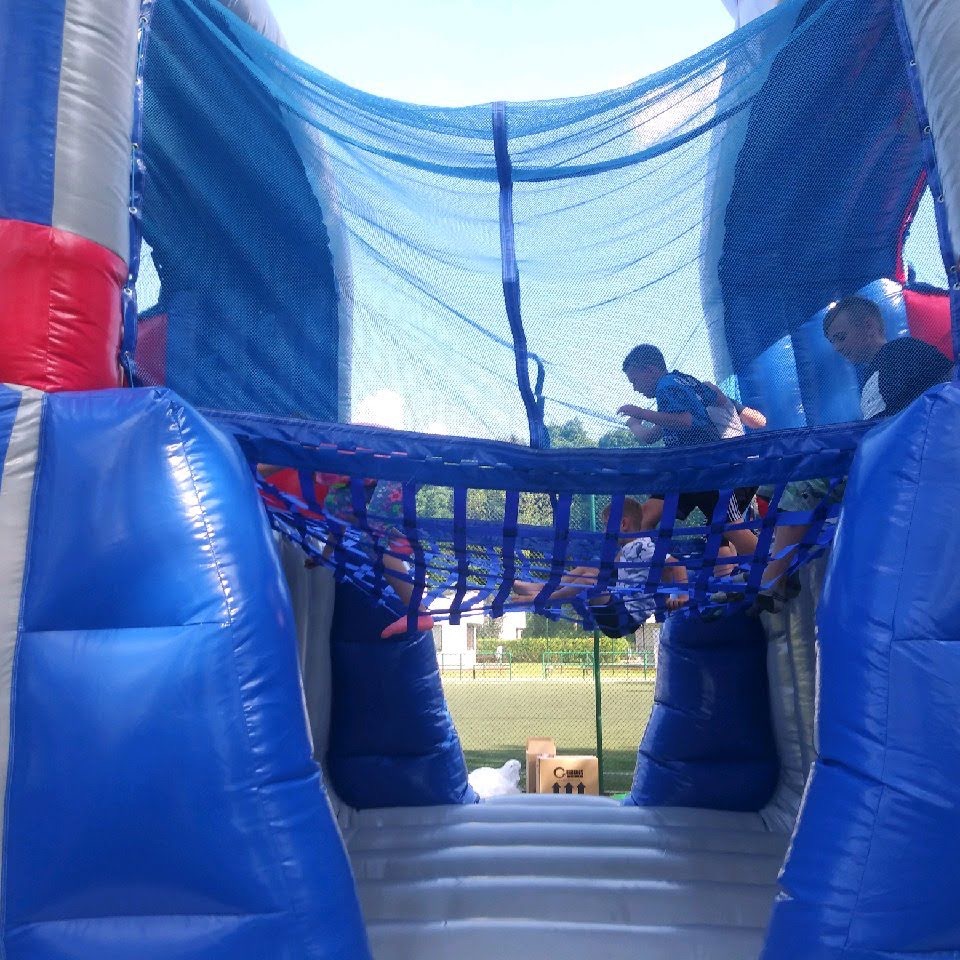 Dzieci bawią się w niebieskim dmuchanym zamku z siatką, widok od wewnątrz, zieleń w tle. Atrakcja eventowa.