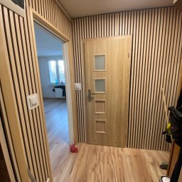 DIM BUD DMYTRO PUKSHYN - Korytarz wykończony panelami ściennymi z drewna z pionowymi czarnymi paskami, drzwi wewnętrzne z czterema przeszkleniami, podłoga laminowana, widok w głąb mieszkania.