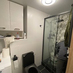 DIM BUD DMYTRO PUKSHYN - Nowoczesna łazienka z czarną armaturą i kabiną prysznicową z wzorzystą ścianą, widoczny fragment osoby wykonującej prace wykończeniowe.