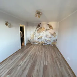 Pusty pokój po remoncie z nową podłogą z paneli w odcieniach brązu, ozdobną tapetą z motywem roślinnym na jednej ścianie i nowoczesnym oświetleniem.