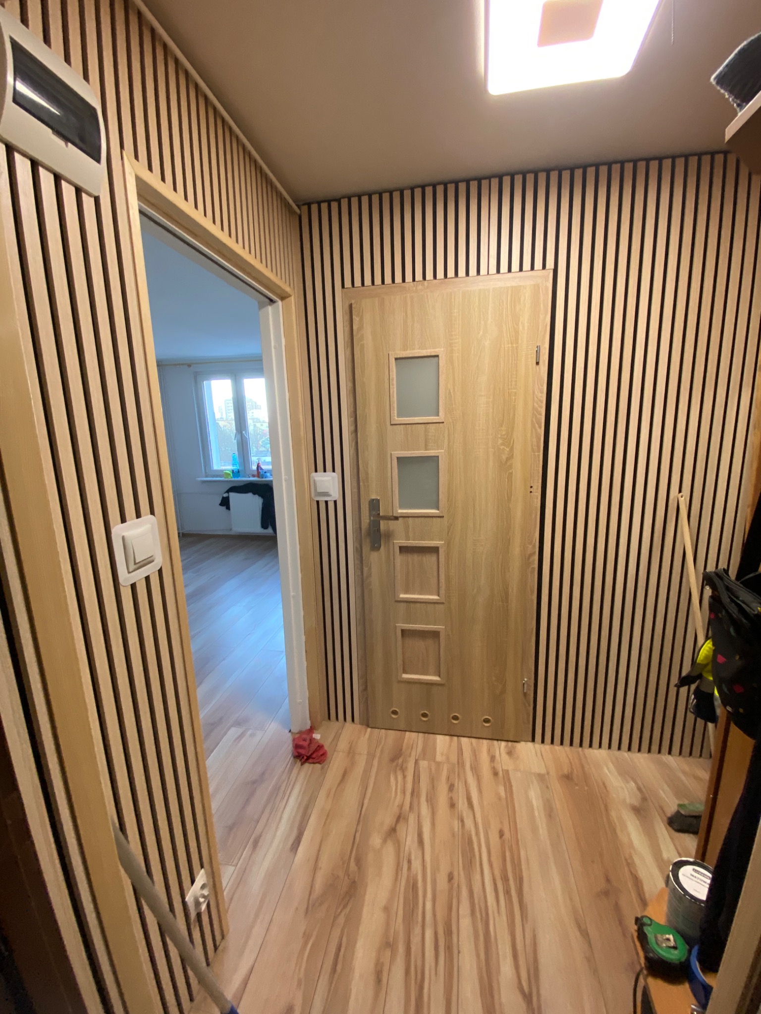 Korytarz wykończony panelami ściennymi z drewna z pionowymi czarnymi paskami, drzwi wewnętrzne z czterema przeszkleniami, podłoga laminowana, widok w głąb mieszkania.