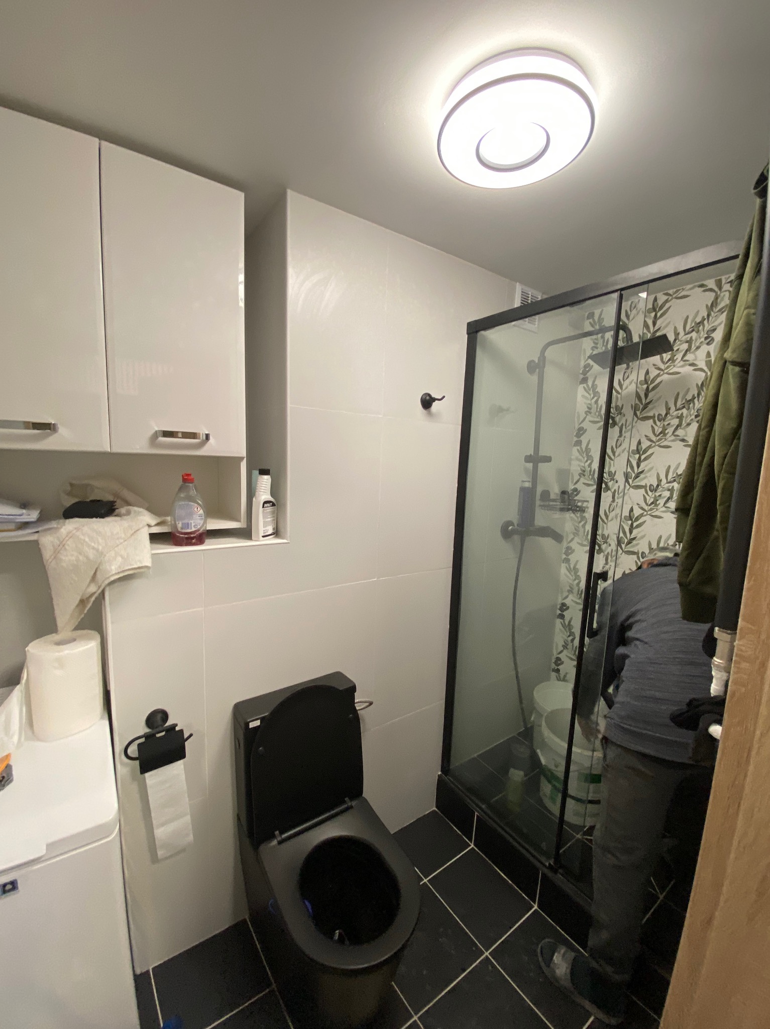 Nowoczesna łazienka z czarną armaturą i kabiną prysznicową z wzorzystą ścianą, widoczny fragment osoby wykonującej prace wykończeniowe.