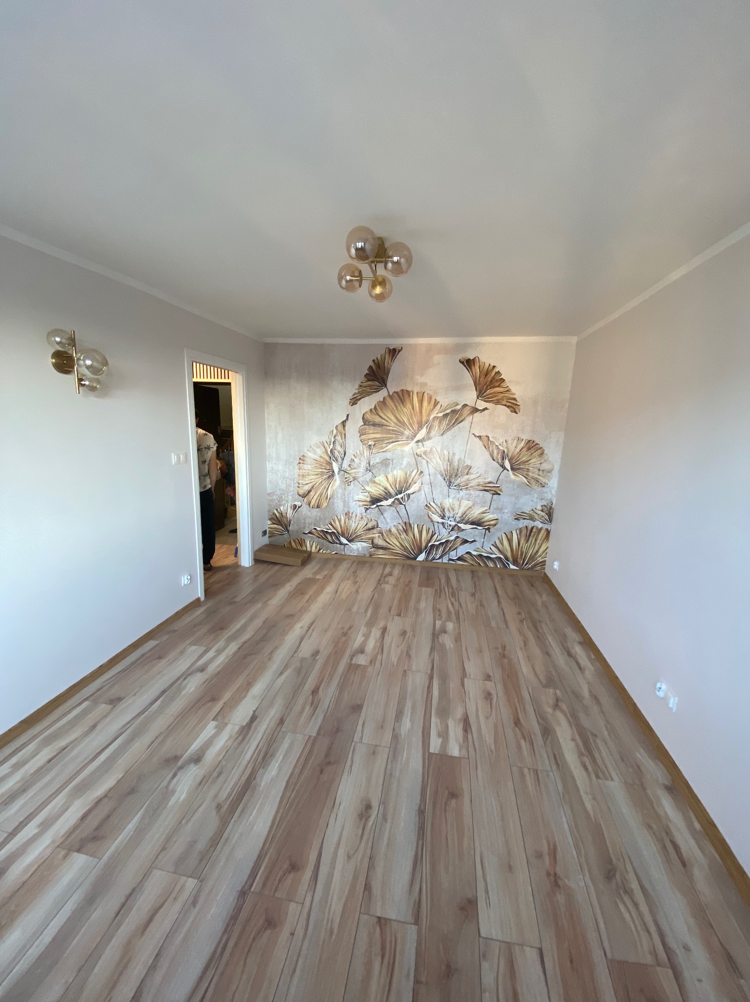 Pusty pokój po remoncie z nową podłogą z paneli w odcieniach brązu, ozdobną tapetą z motywem roślinnym na jednej ścianie i nowoczesnym oświetleniem.