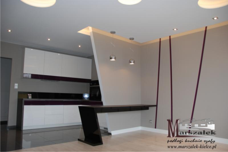 Nowoczesna kuchnia z białymi szafkami, czarnym blatem i elementami dekoracyjnymi w kolorze śliwkowym, oświetlona halogenami i wiszącymi lampami. Widoczny fragment barku z czarnym, błyszczącym...