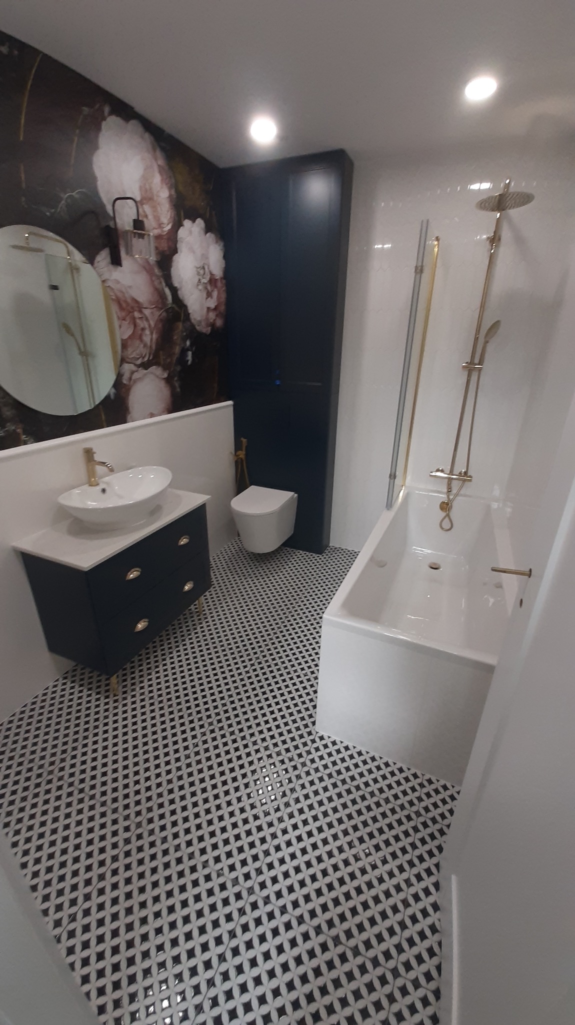 Nowoczesna łazienka z białą armaturą, czarną szafką pod umywalką, złotymi akcentami i wzorzystą czarno-białą podłogą, ściana z motywem kwiatowym.