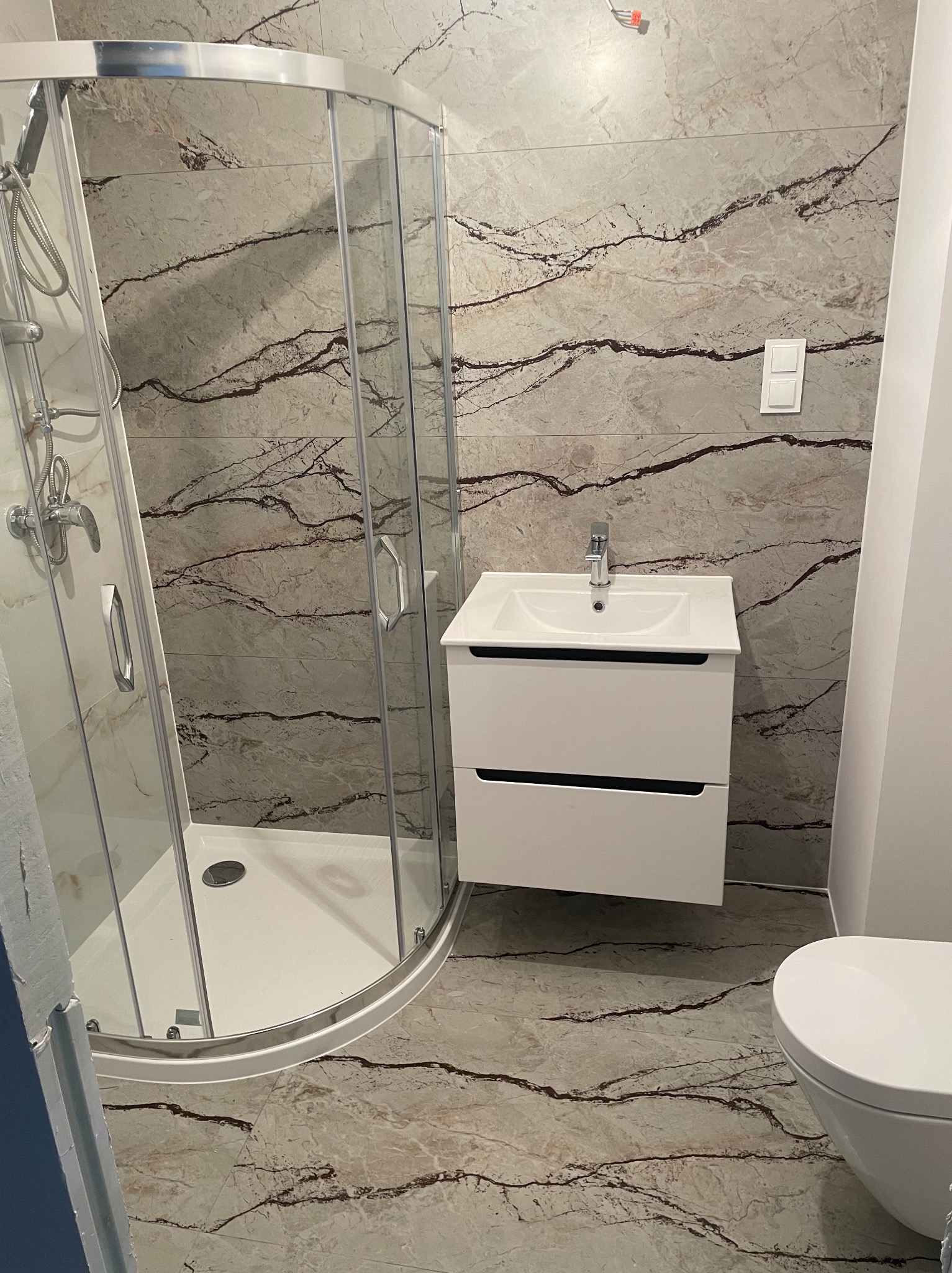 Nowoczesna łazienka z kabiną prysznicową narożną, umywalką z szafką i toaletą, wykończona płytkami imitującymi marmur z ciemnymi żyłami.