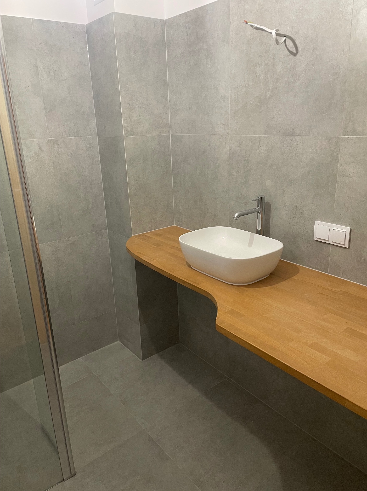 Umywalka nablatowa w kształcie łódki, zamontowana na drewnianym blacie o zaokrąglonym kształcie, szare płytki na ścianach, widoczne przewody elektryczne wystające ze ściany, częściowo widoczna...