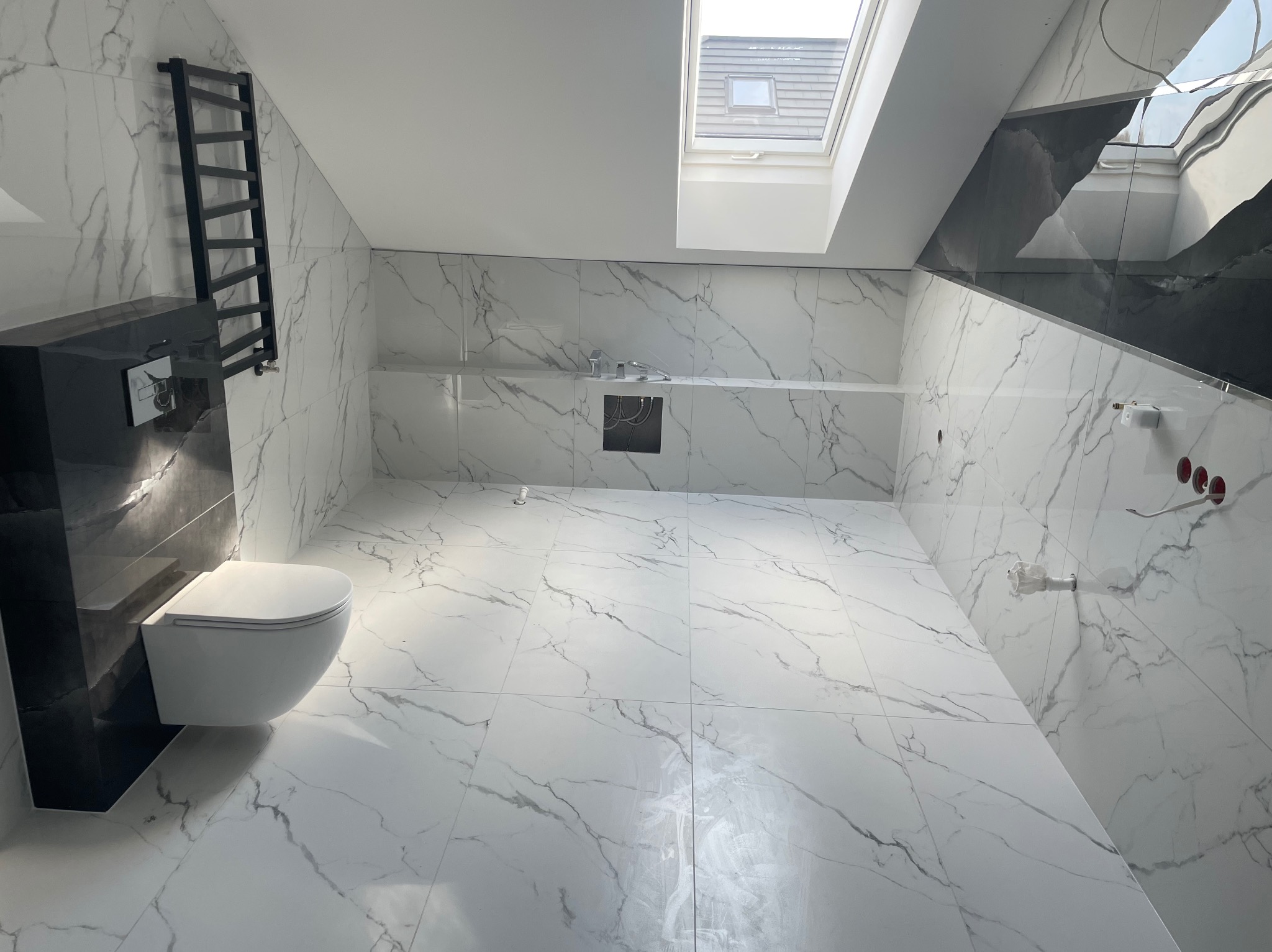 Nowoczesna łazienka na poddaszu z białymi płytkami imitującymi marmur, czarnym grzejnikiem drabinkowym i wiszącą toaletą, doświetlona oknem dachowym.