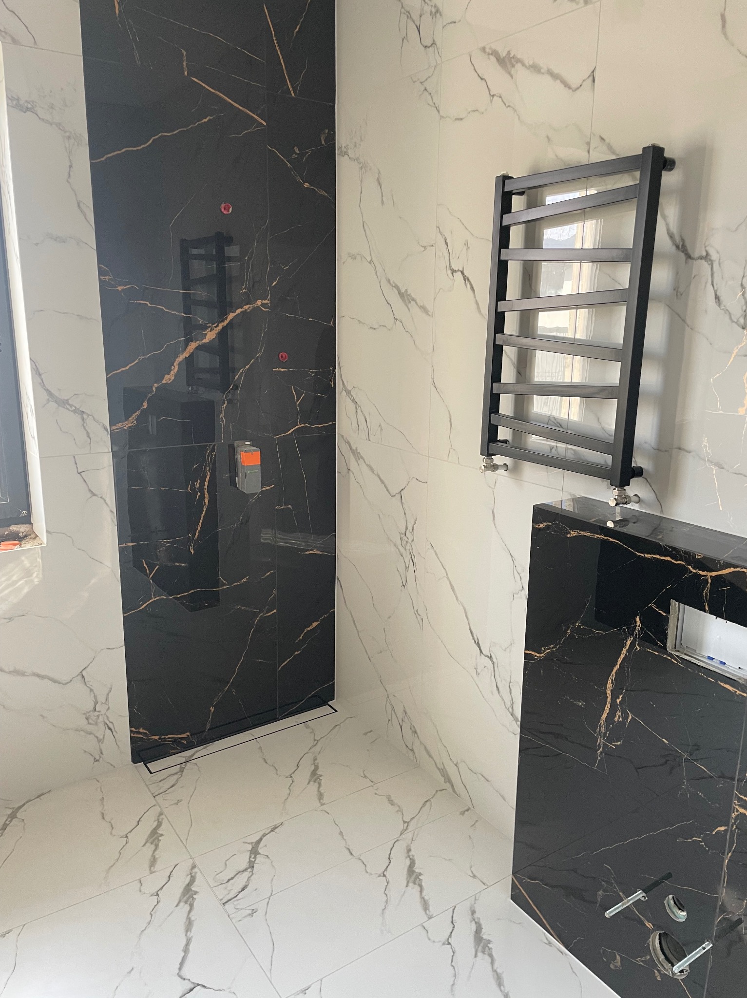 Nowoczesna łazienka z czarnymi i białymi płytkami imitującymi marmur, czarny grzejnik drabinkowy i zabudowa umywalki, widoczne elementy instalacji.