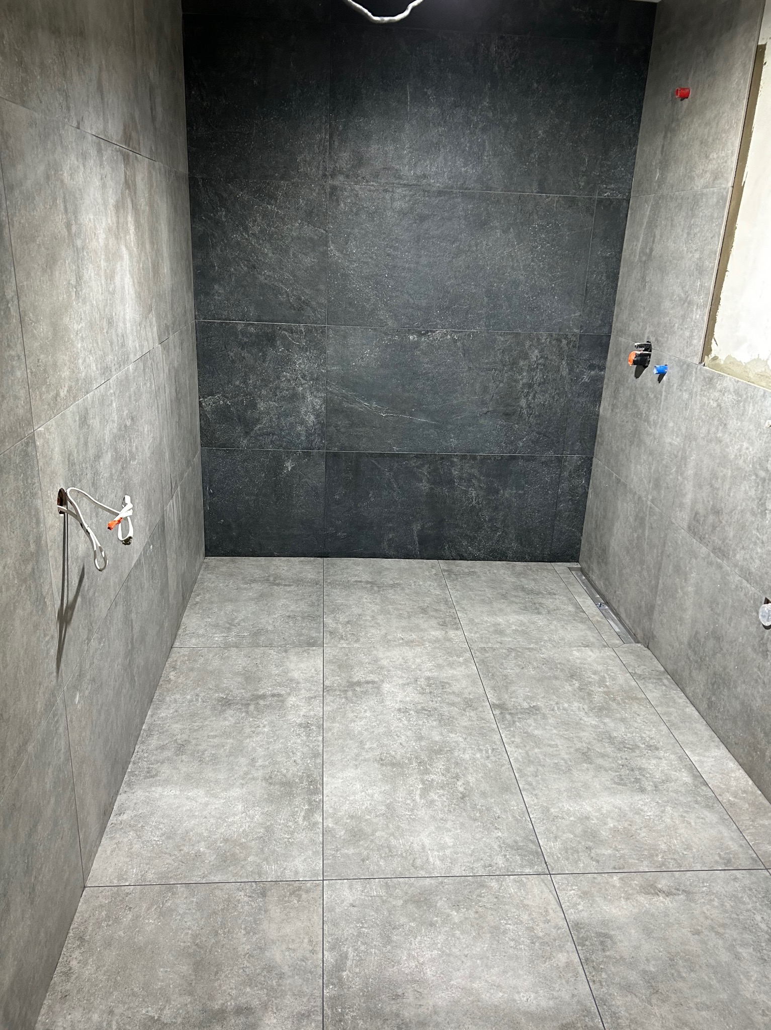 Wnętrze łazienki w trakcie prac wykończeniowych, szare płytki na ścianach i podłodze, widoczne przyłącza wodne i elektryczne, fragment niezakończonej ściany z widocznym tynkiem.