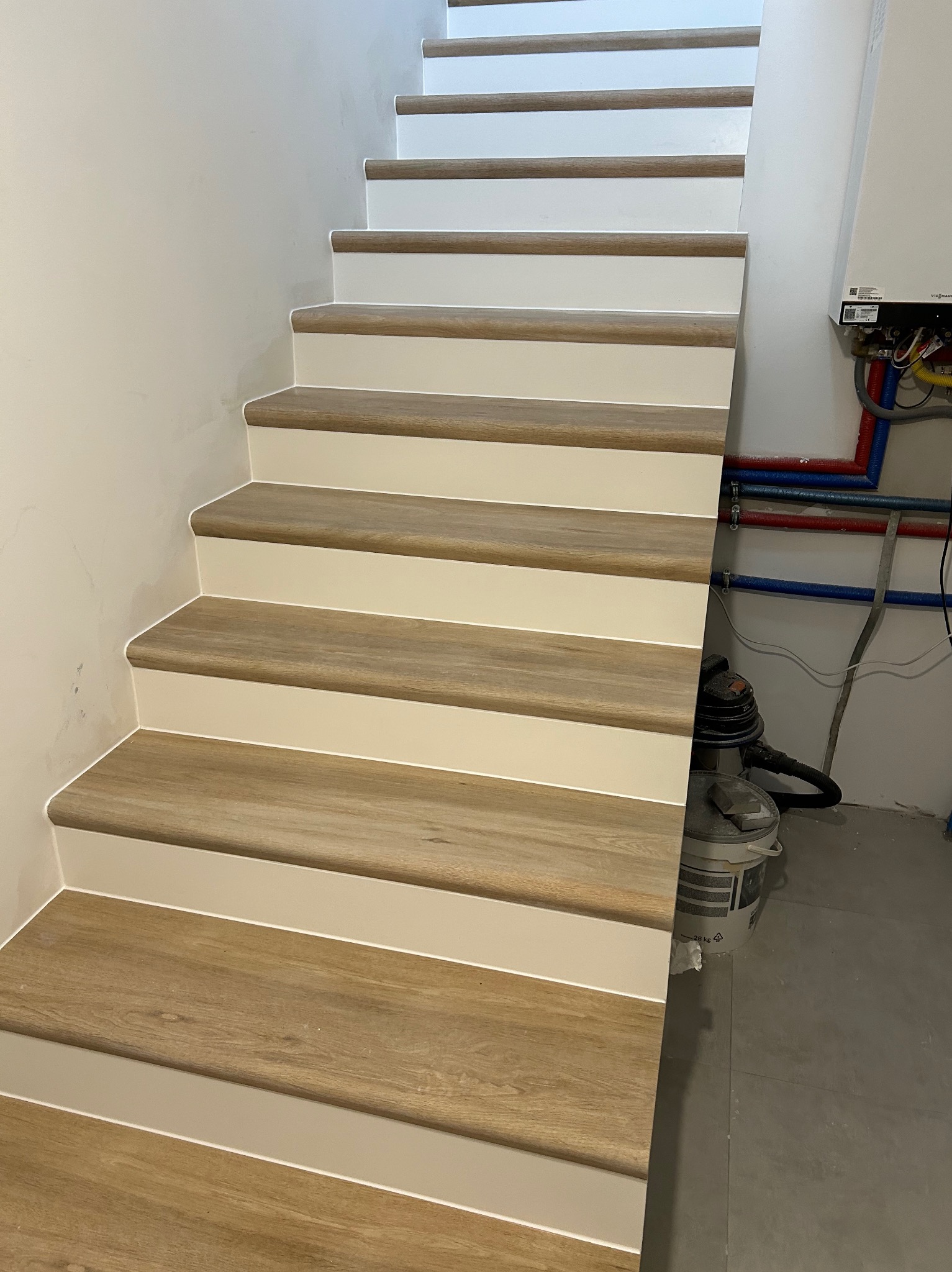 Schody z drewnopodobnymi stopniami i białymi podstopnicami, widoczny odkurzacz i piec gazowy w tle.