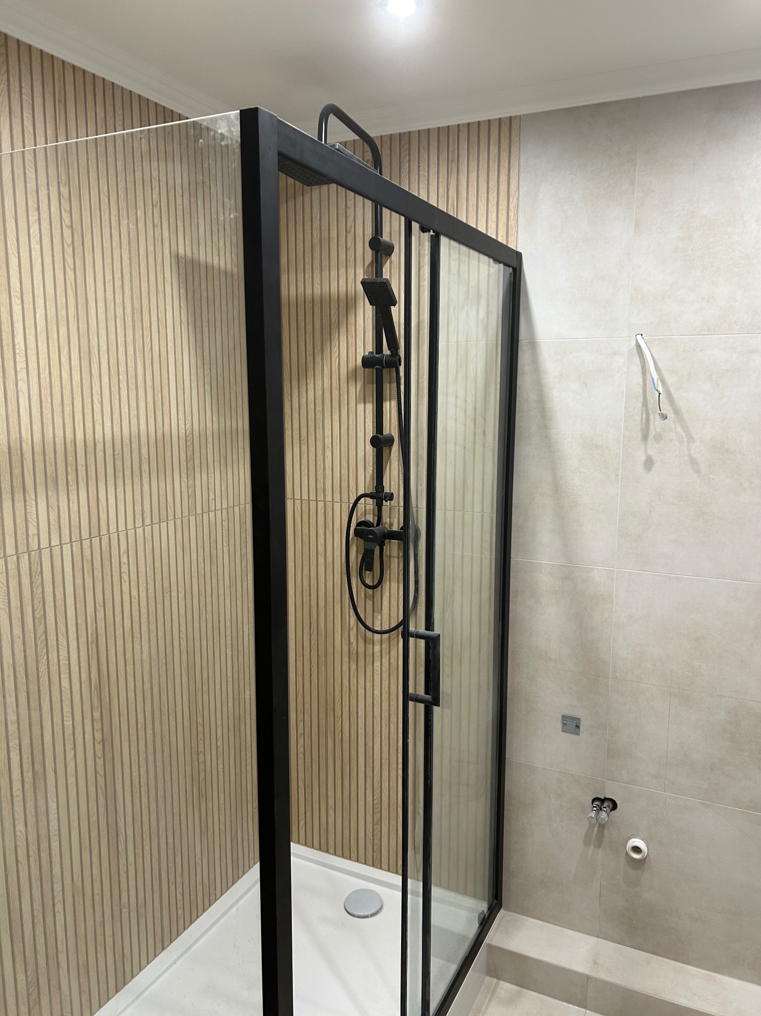 Nowoczesna kabina prysznicowa z czarną ramą i deszczownicą, zamontowana w łazience wykończonej płytkami imitującymi drewno i beżowymi płytkami.