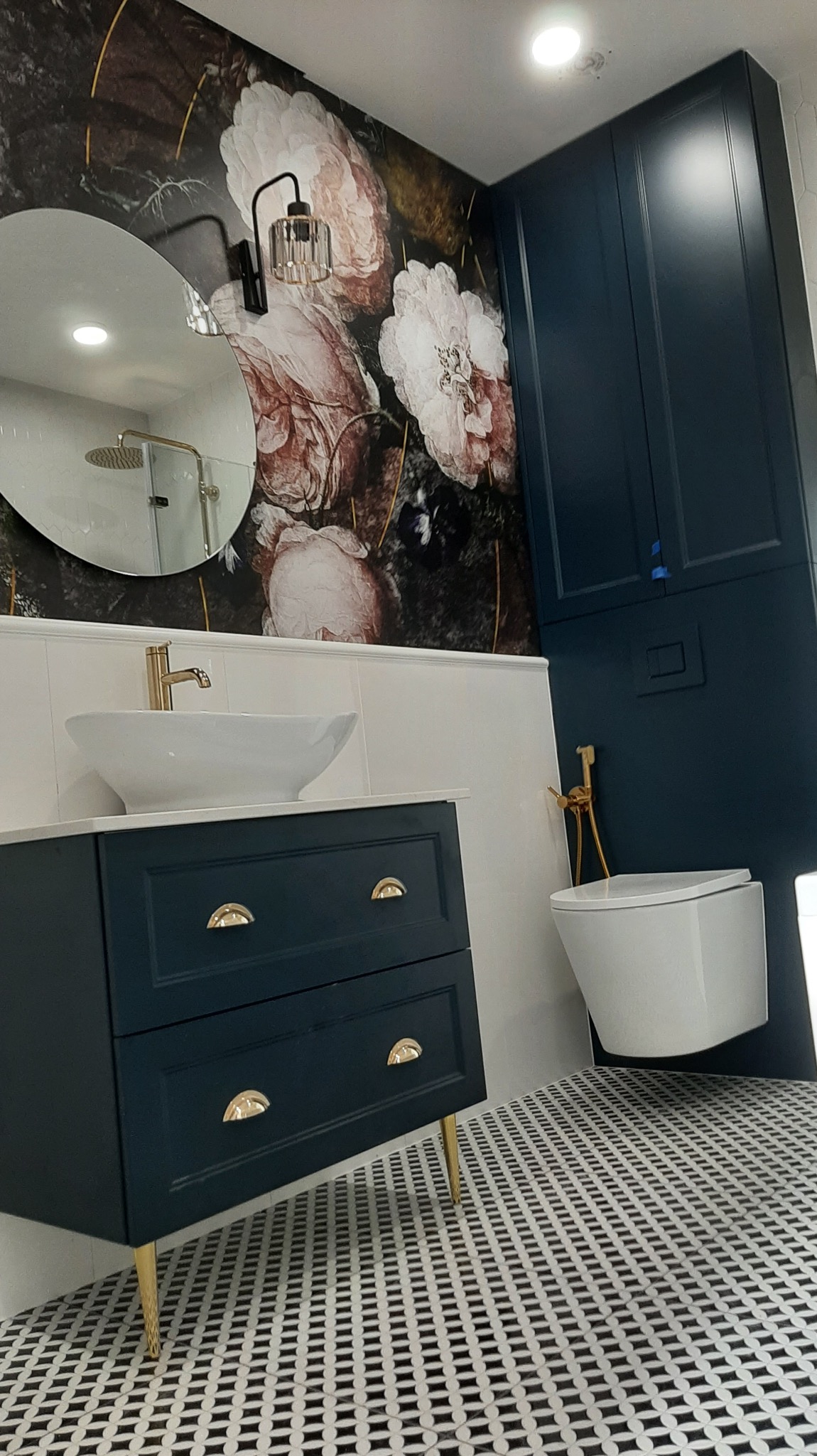 Nowoczesna łazienka z ciemną szafką pod umywalkę na złotych nóżkach, owalnym lustrem, toaletą podwieszaną i wzorzystą podłogą w czarno-białe romby na tle ściany z tapetą w kwiaty.