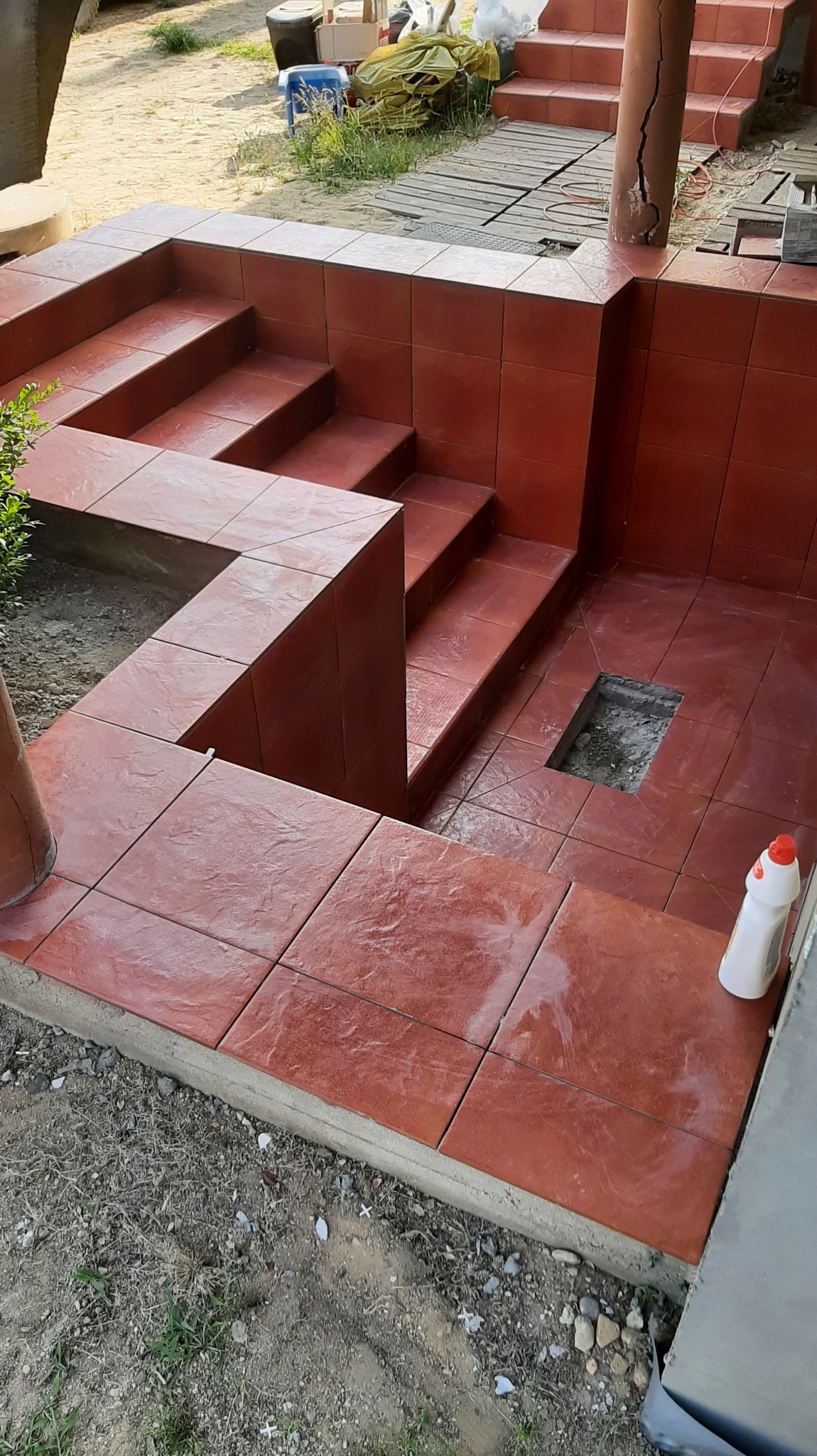 Czerwone płytki podłogowe i schodowe na zewnątrz, z widocznym otworem w podłodze i butelką płynu czyszczącego.