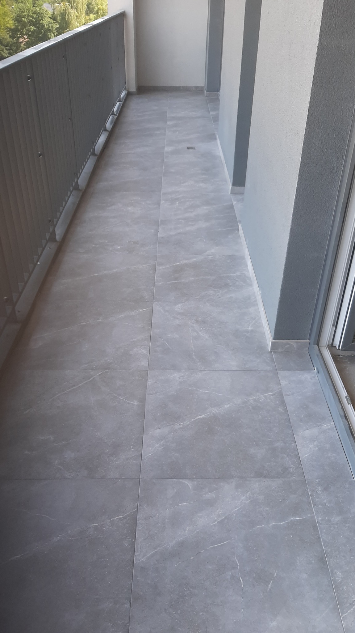 Wyłożony szarymi płytkami balkon z metalową barierką i odpływem liniowym w posadzce.