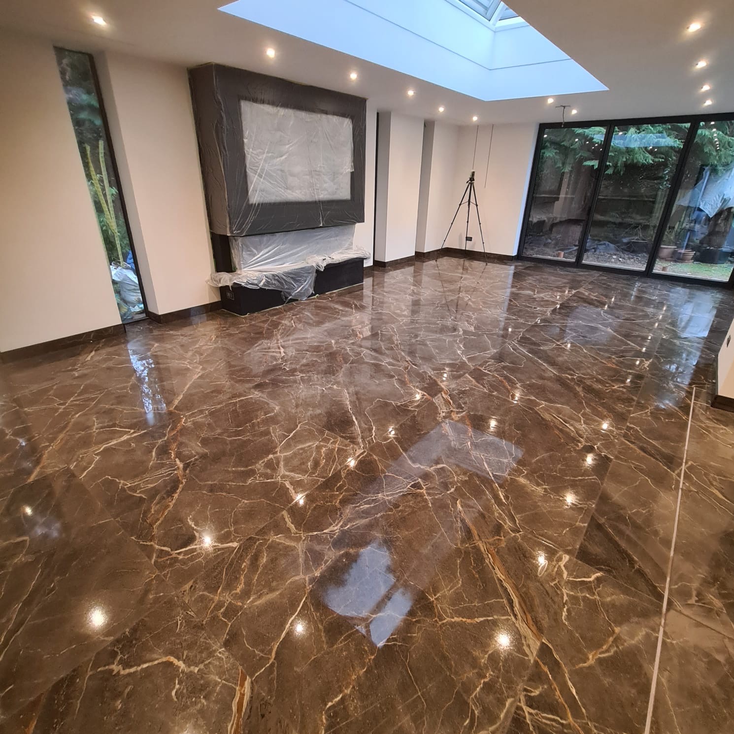 Nowoczesne wnętrze z błyszczącą, brązową podłogą z płytek imitujących marmur, osłoniętym folią kominkiem, dużymi oknami z widokiem na zieleń ogrodu oraz oknem dachowym w suficie.