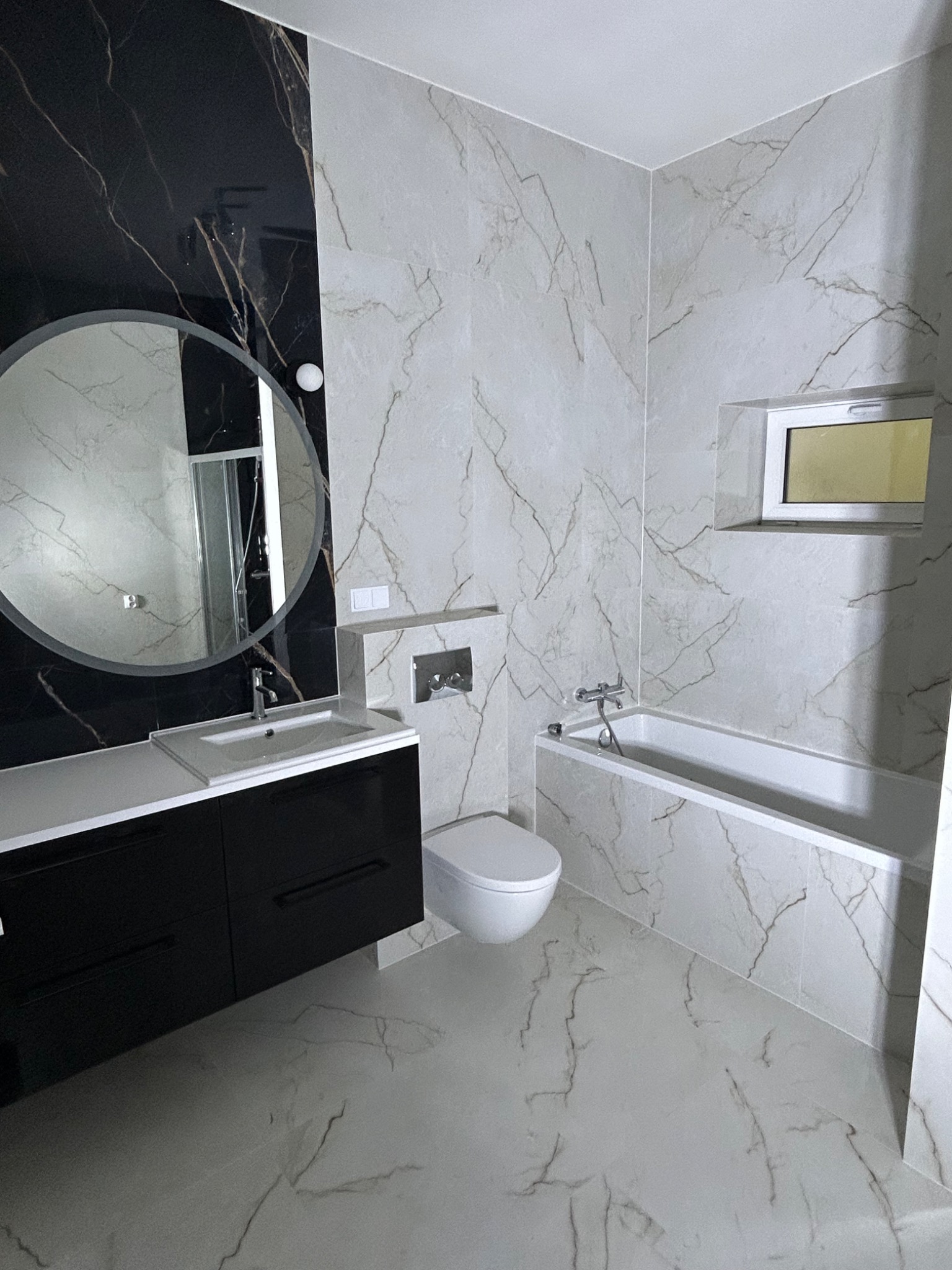 Elegancka łazienka z czarno-białymi płytkami marmurkowymi, okrągłym lustrem nad czarną szafką umywalkową i białą wanną. Minimalistyczny styl, nowoczesne wykończenie.