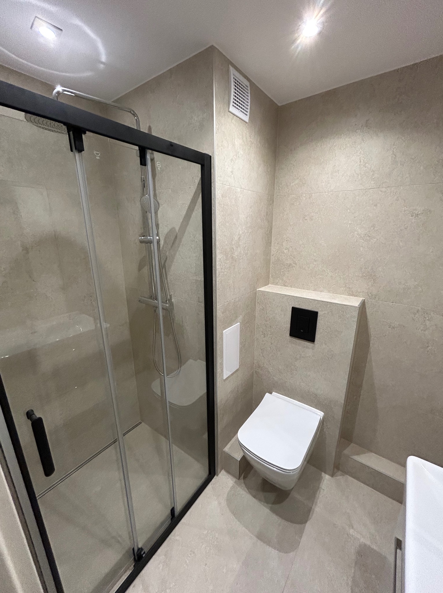 Jasna łazienka po remoncie: kabina prysznicowa z czarnymi ramami, podwieszana toaleta z zabudową, płytki w odcieniach beżu i szarości, minimalistyczny design.
