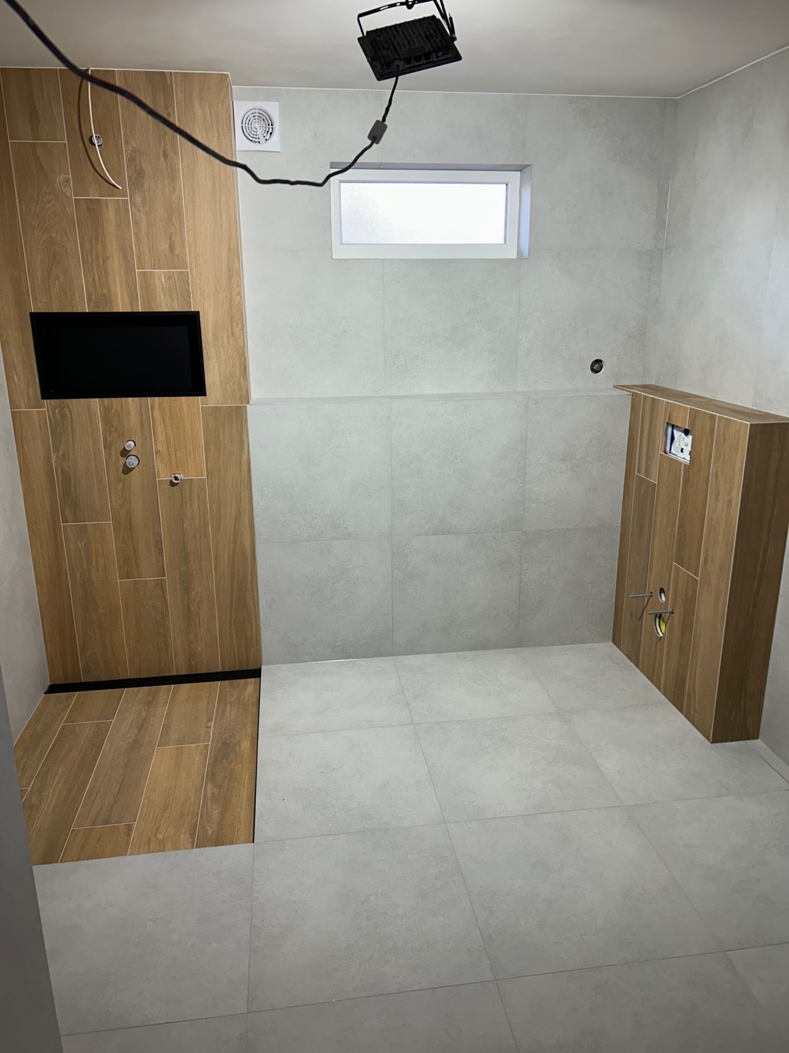 Nowoczesna łazienka w Białymstoku z drewnopodobnymi i szarymi płytkami. Widoczna zabudowa stelaża WC i wnęka na TV. Oświetlenie robocze na suficie.