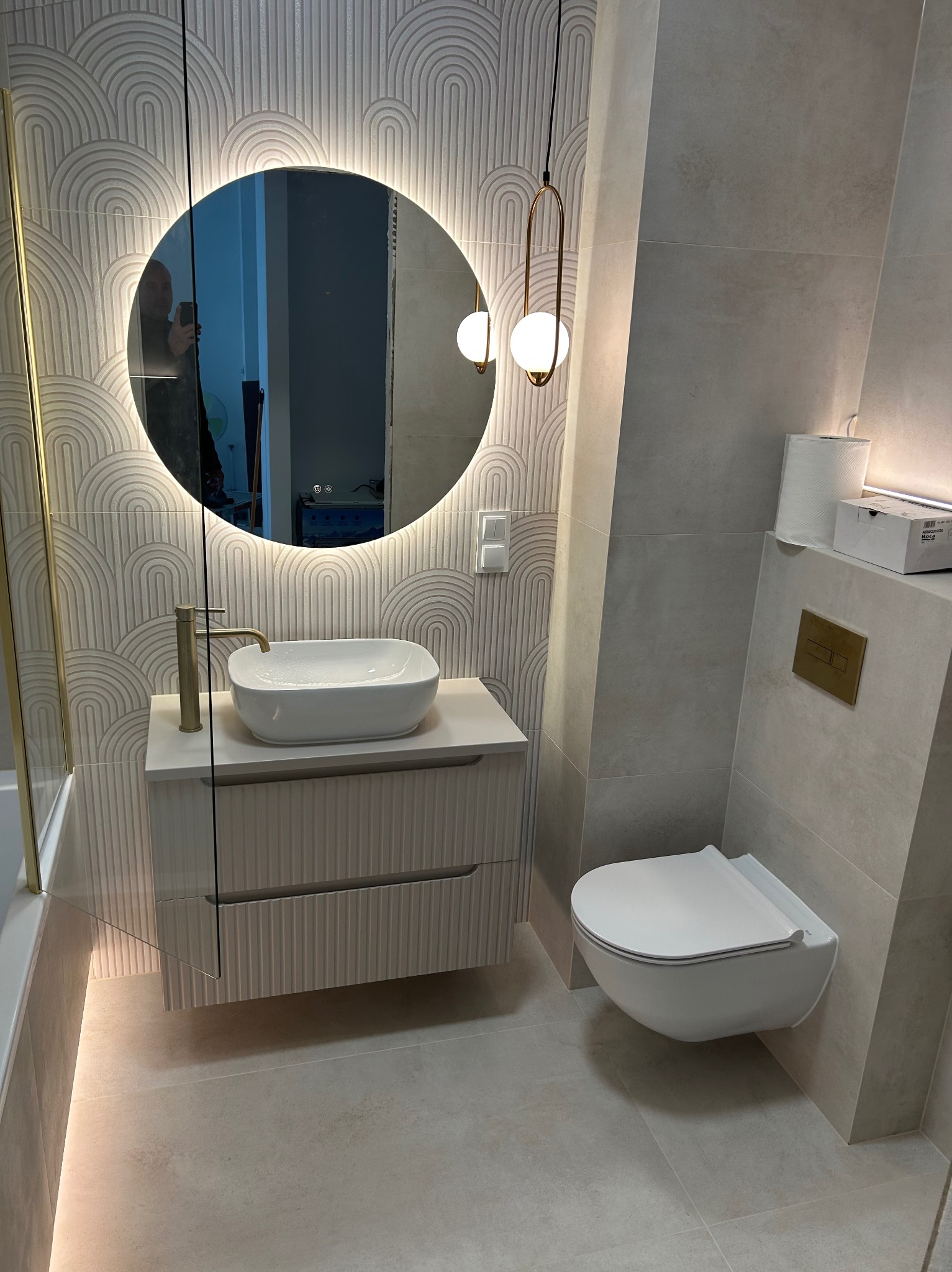 Nowoczesna łazienka z podświetlanym lustrem, wiszącą toaletą i umywalką na szafce z ryflowanym frontem. Minimalistyczny design, stonowana kolorystyka.