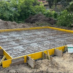 Korz-bud - Zbrojenie fundamentu z żółtym szalunkiem na gruncie, widok z góry. Na pierwszym planie wiadro z zaprawą. W tle zieleń drzew i krzewów.