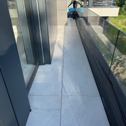 Korz-bud - Nowoczesny balkon z szarą, kwadratową płytką w Warszawie. Balustrada szklana, widok na zieleń. Częściowo zabezpieczony folią, przygotowany do prac wykończeniowych.