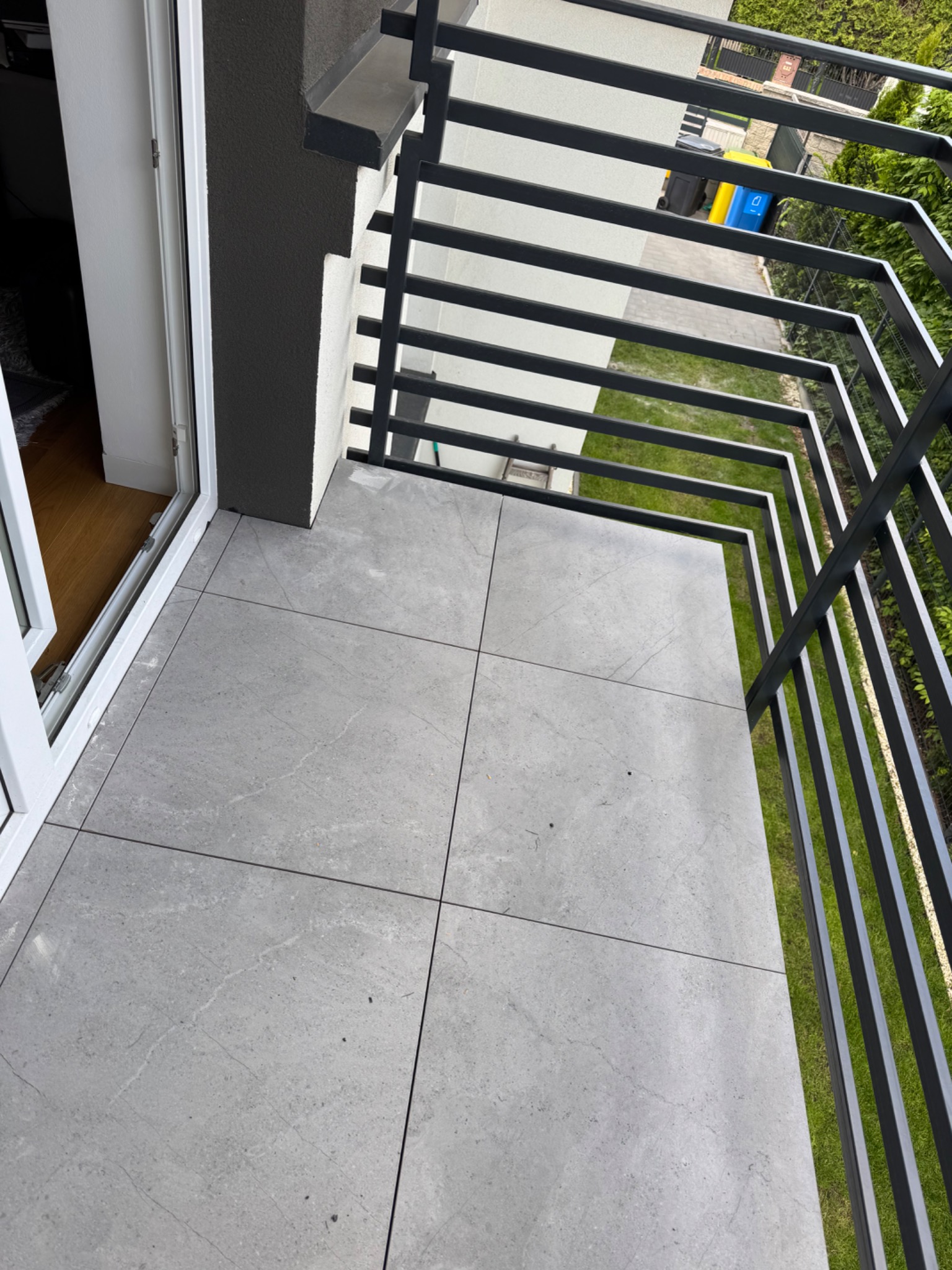 Wykończony balkon z szarymi płytkami i nowoczesną, czarną balustradą. Widok z góry, z otwartymi drzwiami balkonowymi. Na zewnątrz zieleń i elementy otoczenia.