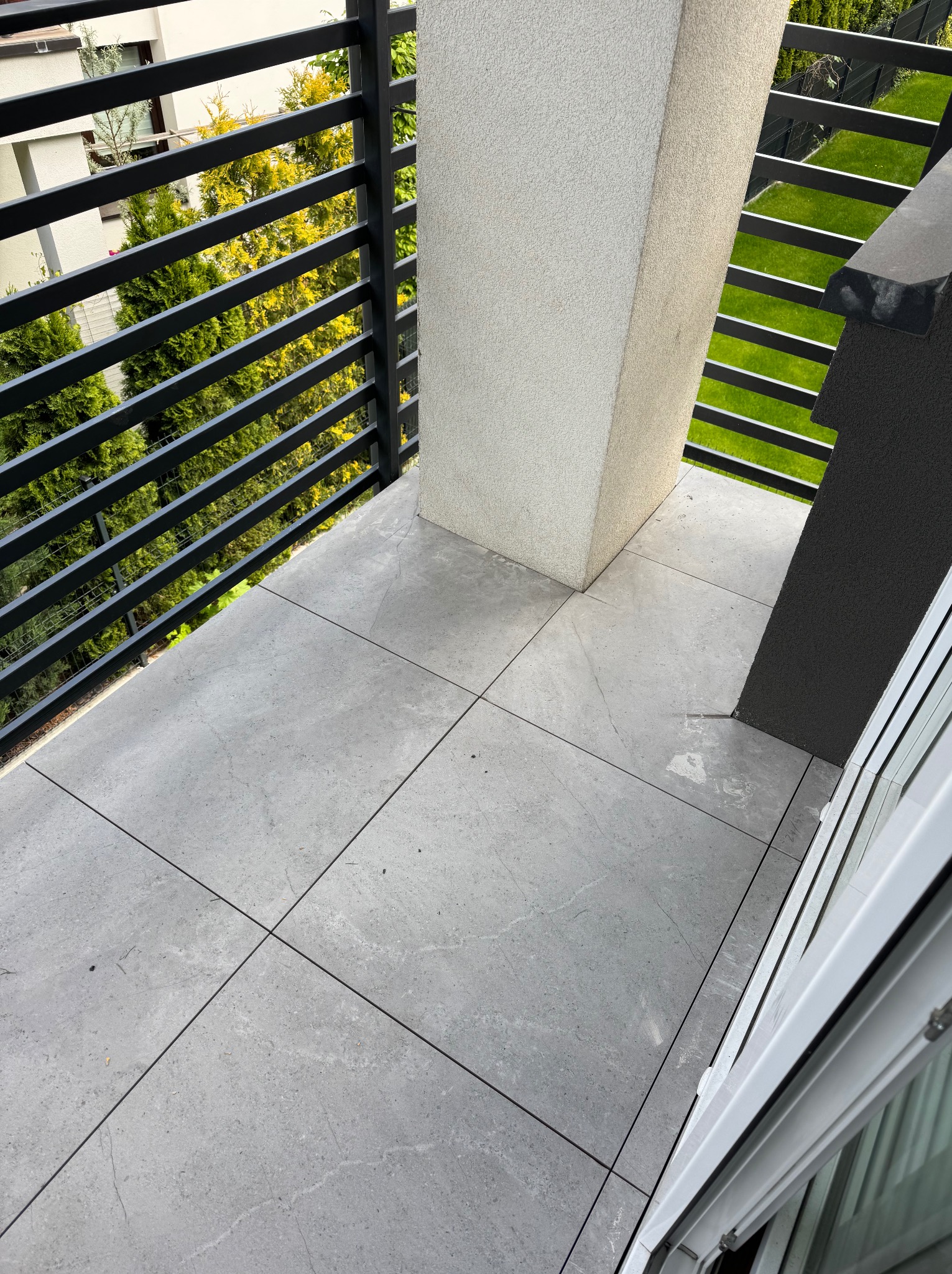 Wykończony balkon z szarą, gładką podłogą z płytek. Czarne, poziome barierki. Widok na zieleń za barierkami. Jasne, naturalne światło.