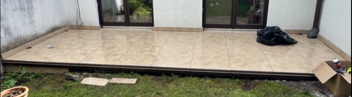 Wykończony taras z beżowych płytek, widoczne drzwi balkonowe i fragment trawnika. Na tarasie leży czarna torba i karton z narzędziami.