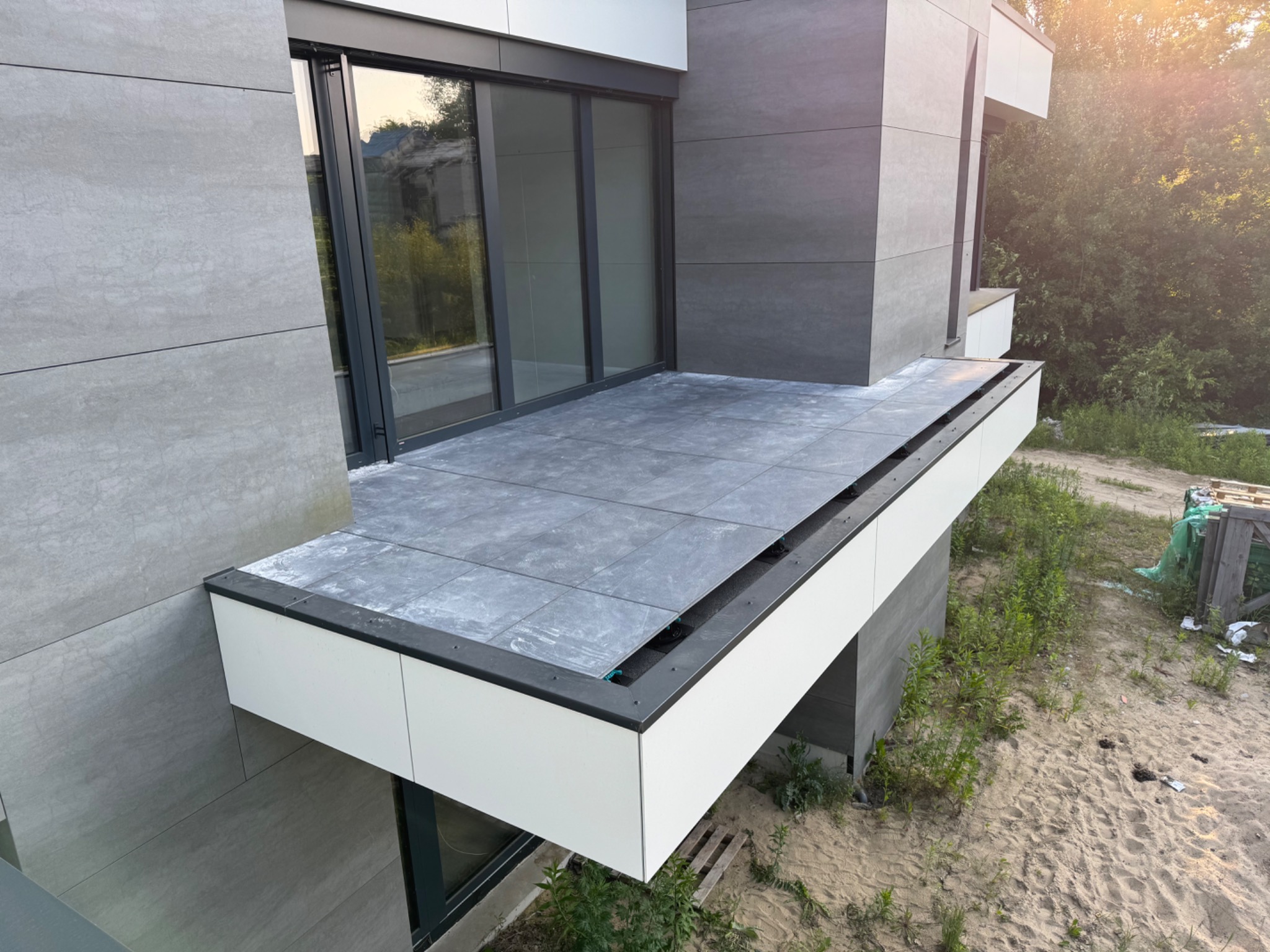 Nowoczesny balkon wykończony szarą płytką, z widocznymi elementami konstrukcyjnymi i minimalistyczną balustradą. Widok z góry na tle zieleni i piasku.