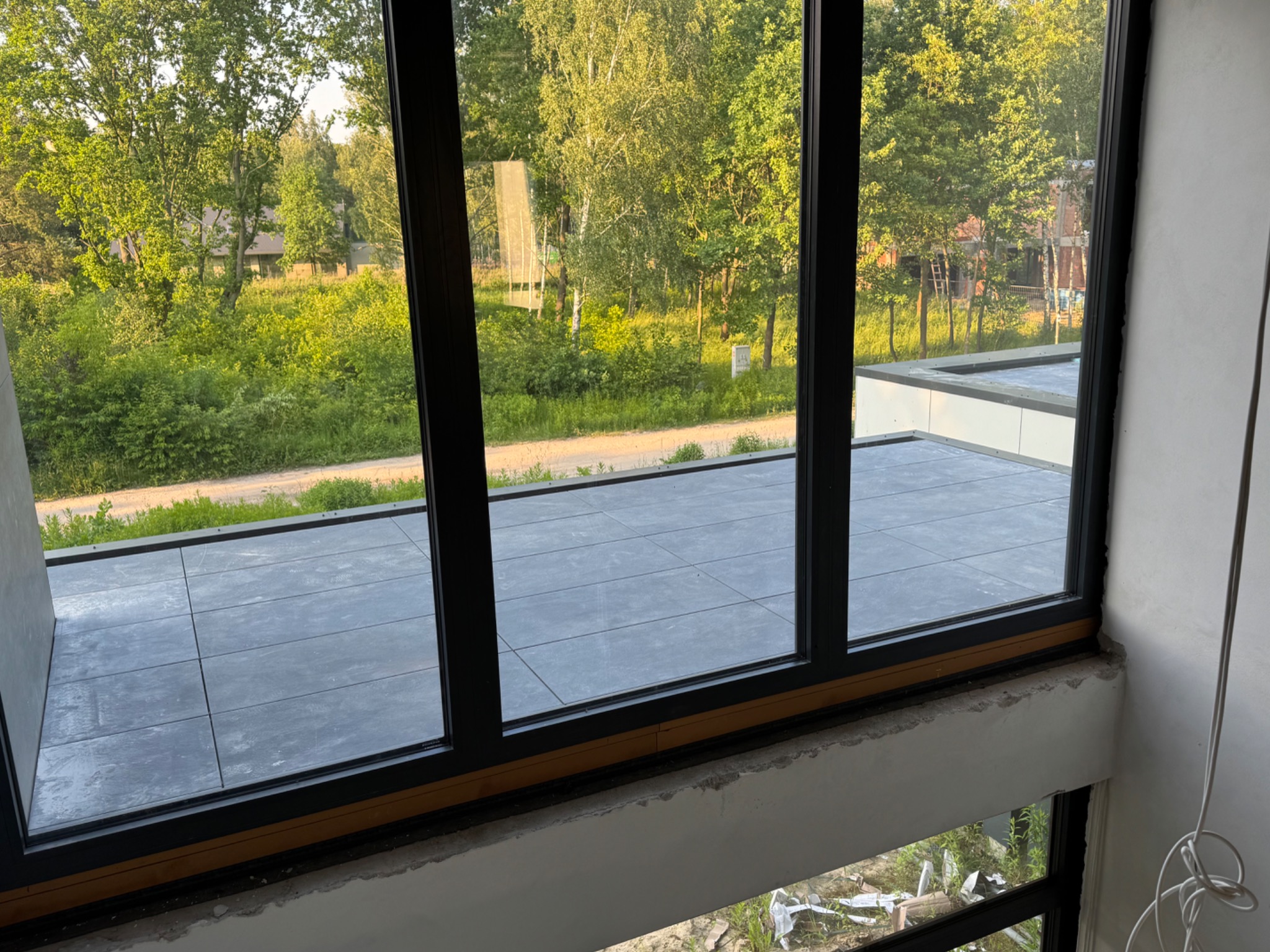 Wykończony taras z szarymi płytkami, widok przez okno na zieleń. Nowoczesny design, widoczne elementy wykończeniowe. Jasne światło dzienne.