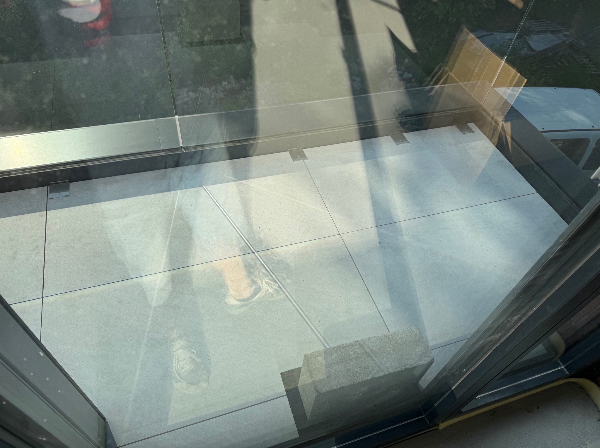 Wykończony balkon z szarymi płytkami i szklaną balustradą, widok z góry przez szybę, z odbiciem osoby. Minimalistyczny design.
